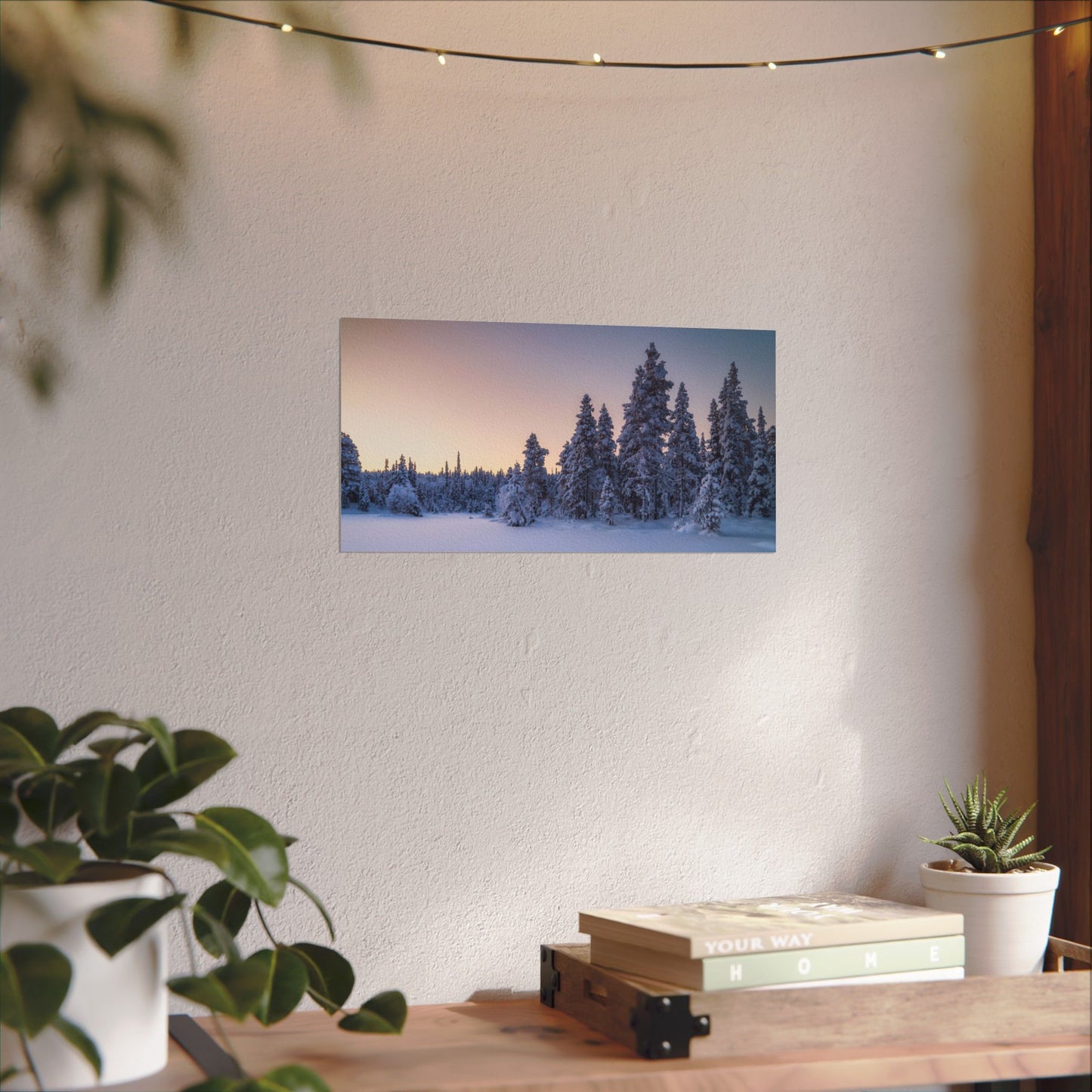 Framed winter landscape print on a wall above a wooden table with plants.