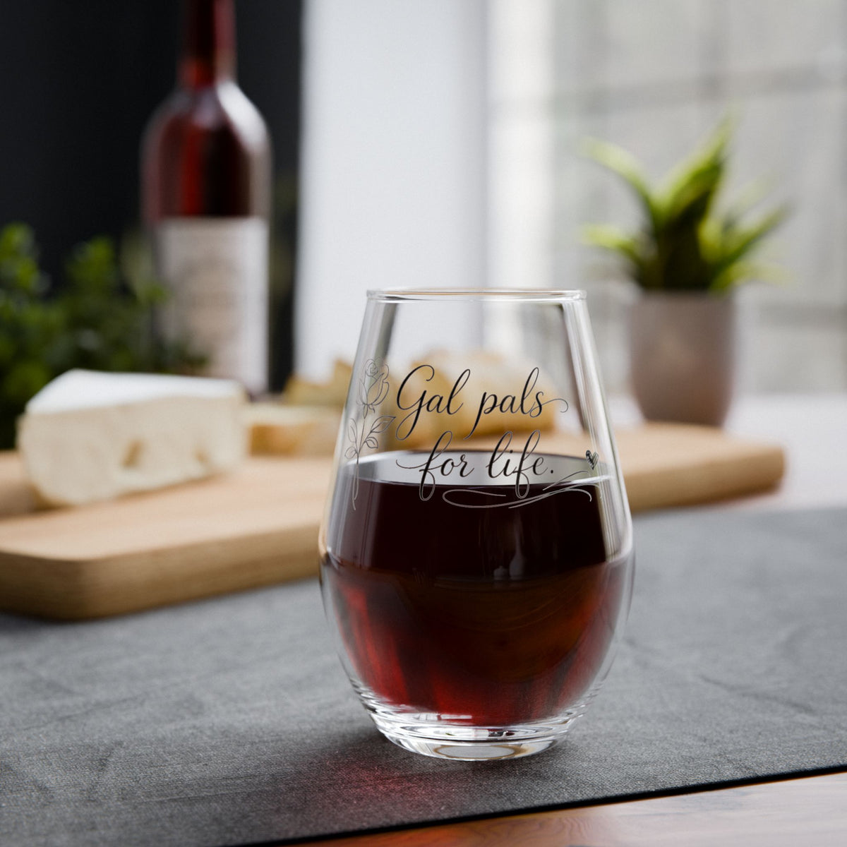 Glass of red wine with 'Gal pals for life' text on a table with cheese and wine bottle in the background.