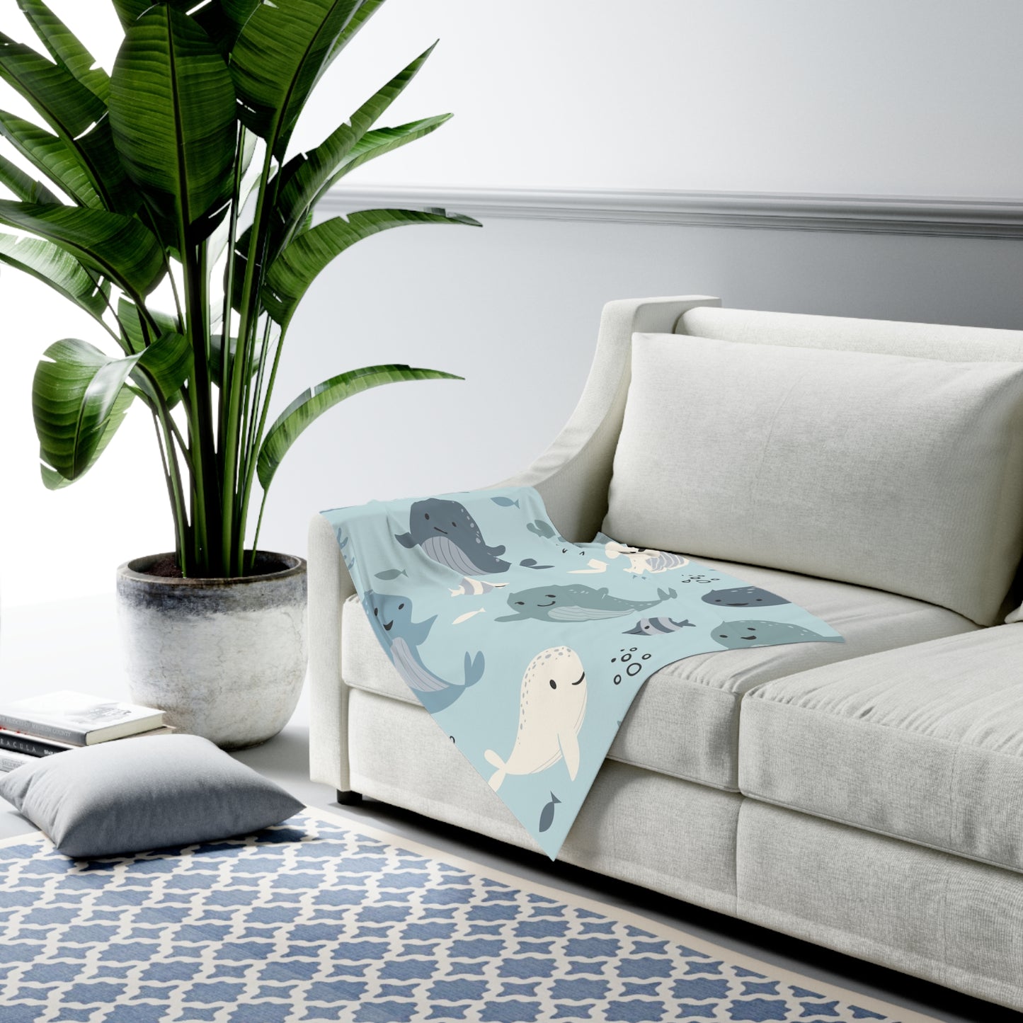 Light gray sofa with a blue blanket featuring whale patterns, next to a potted plant on a white wall.
