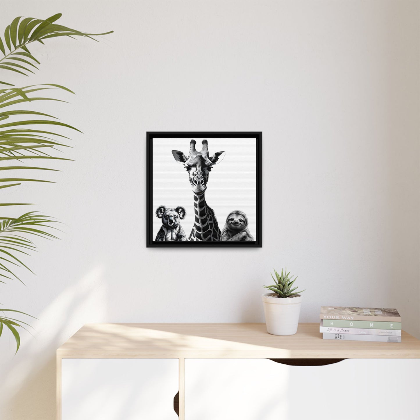 Framed artwork of a giraffe and two animals on a wall above a desk with a plant and books.
