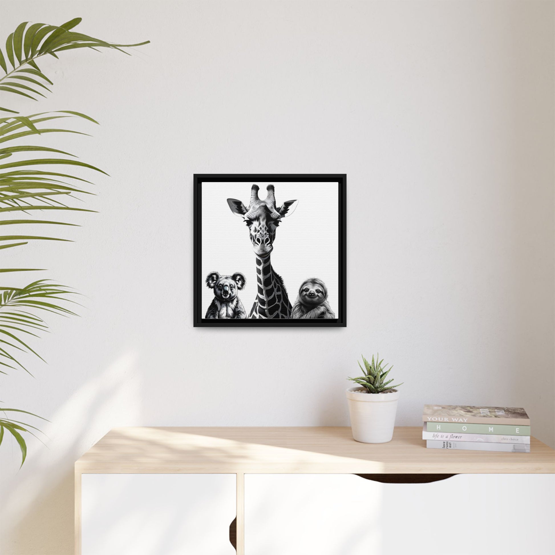 Framed artwork of a giraffe and two animals on a wall above a desk with a plant and books.