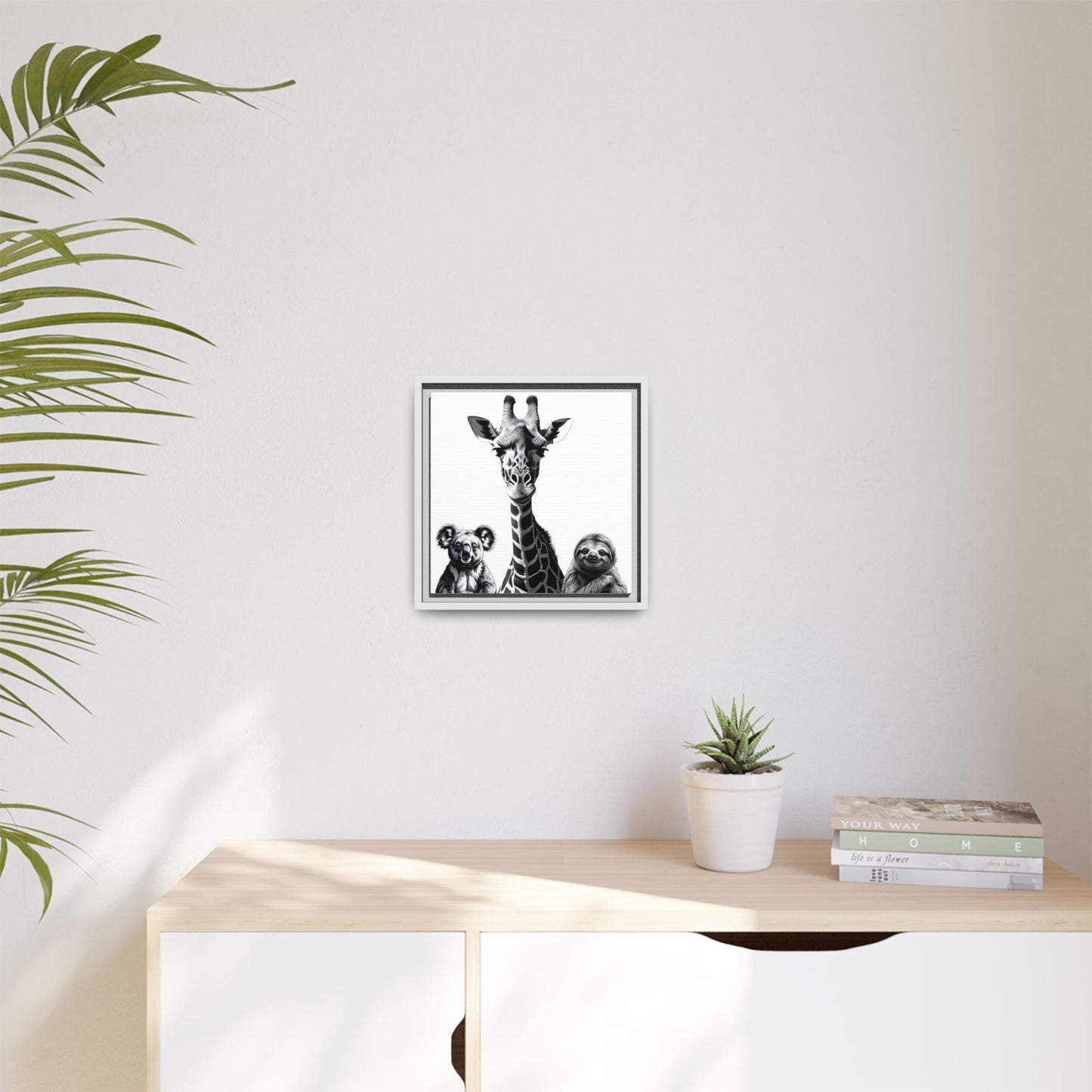 Framed artwork of a giraffe and sloths on a wall above a desk with a plant and books.