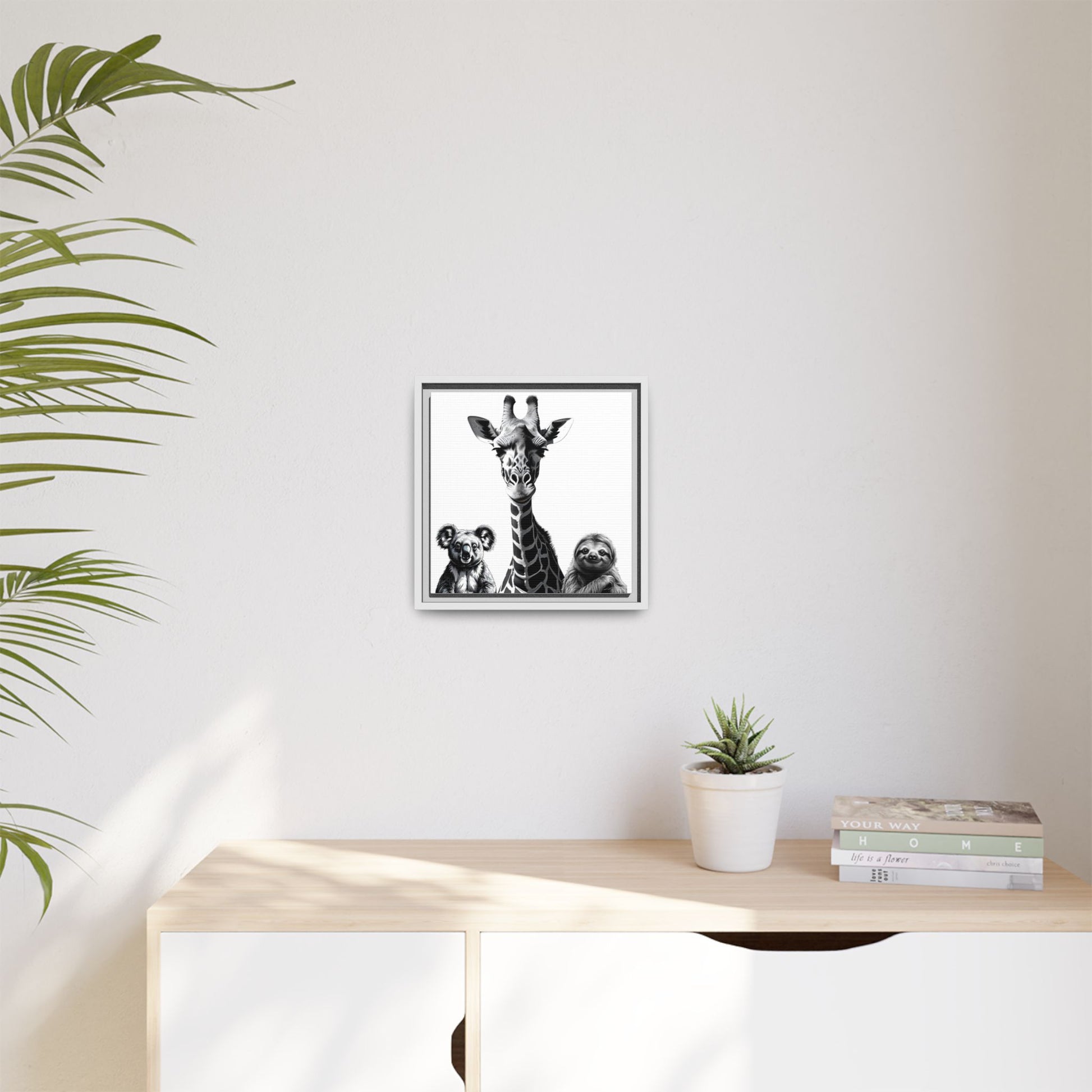 Framed artwork of a giraffe and sloths on a wall above a desk with a plant and books.