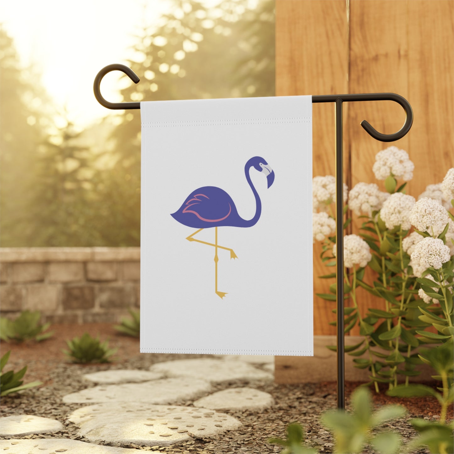 Flamingo-themed garden flag on a stand with flowers and a wooden fence in the background