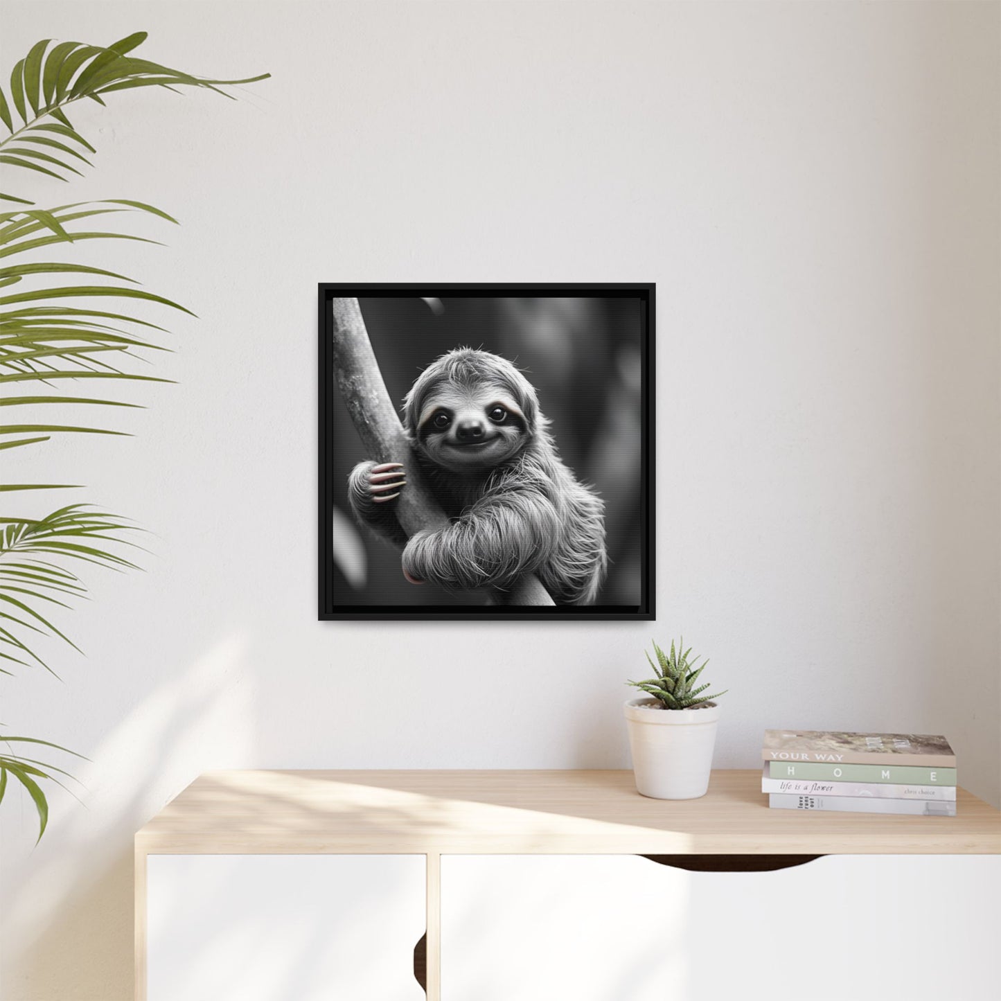 Framed black and white photo of a sloth on a wall above a desk with a plant and books.