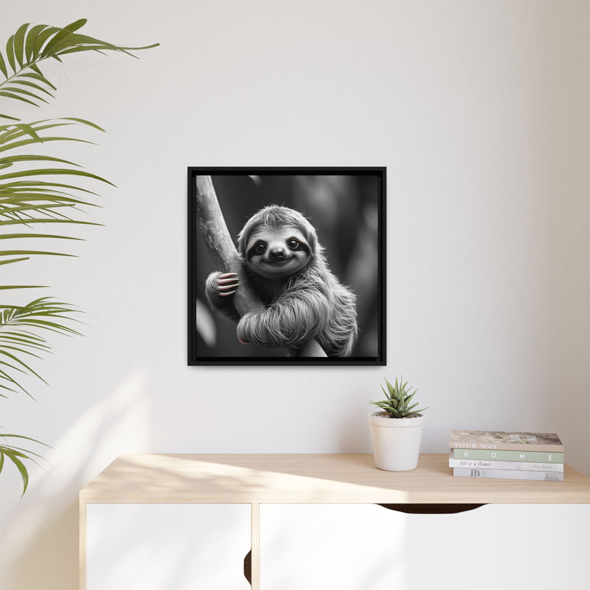 Framed black and white photo of a sloth on a wall above a desk with a plant and books.