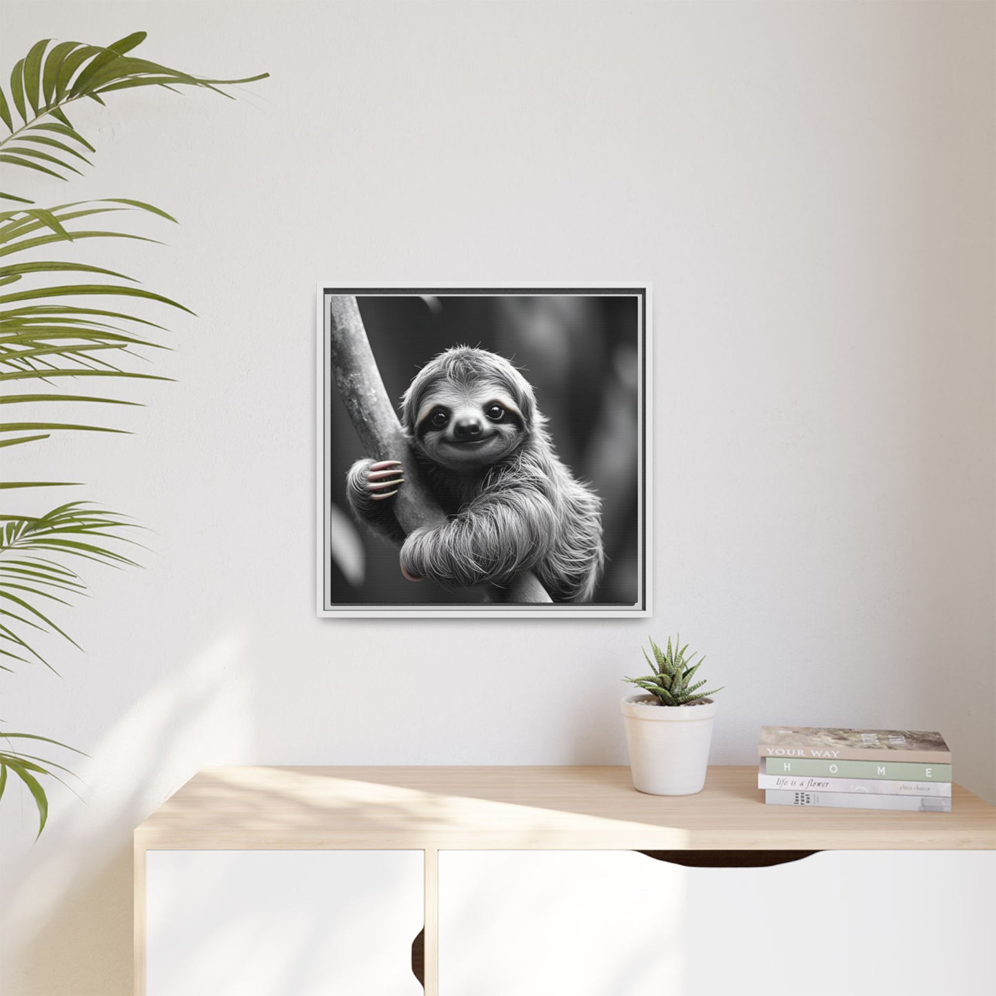 Framed black and white sloth print on a wall above a wooden desk with a plant and books.