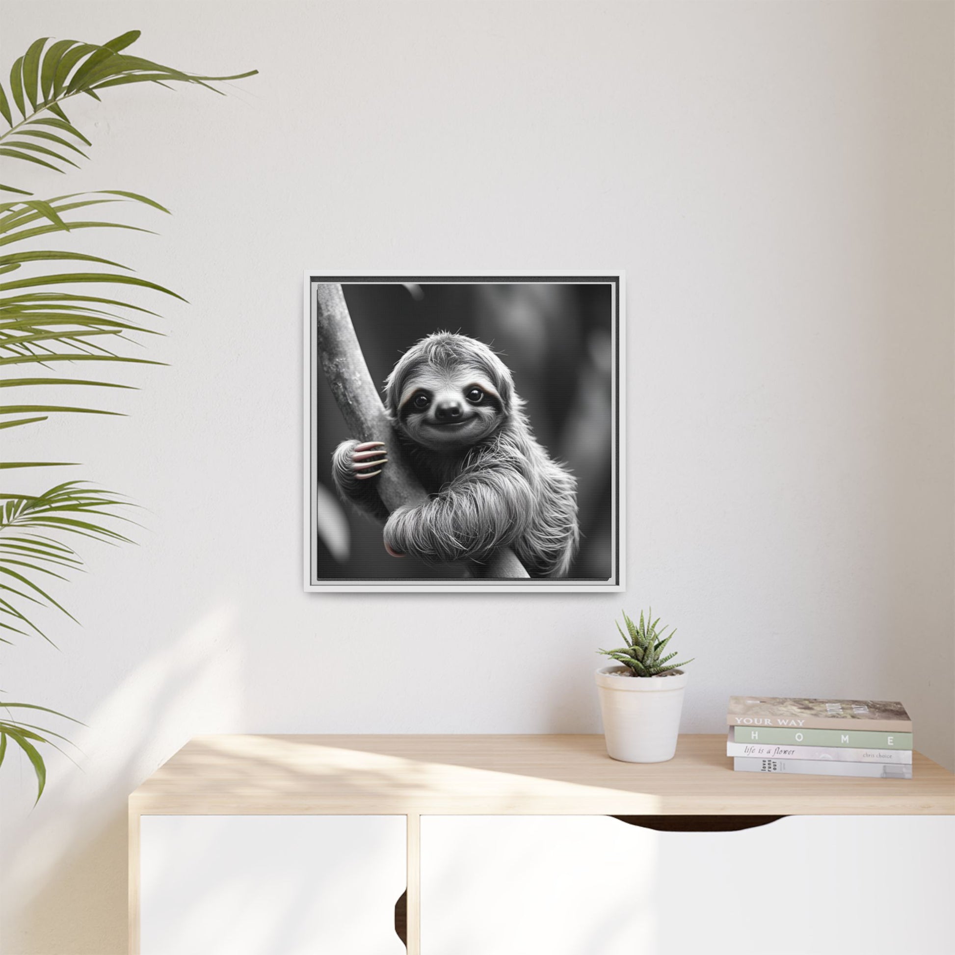 Framed black and white sloth print on a wall above a wooden desk with a plant and books.
