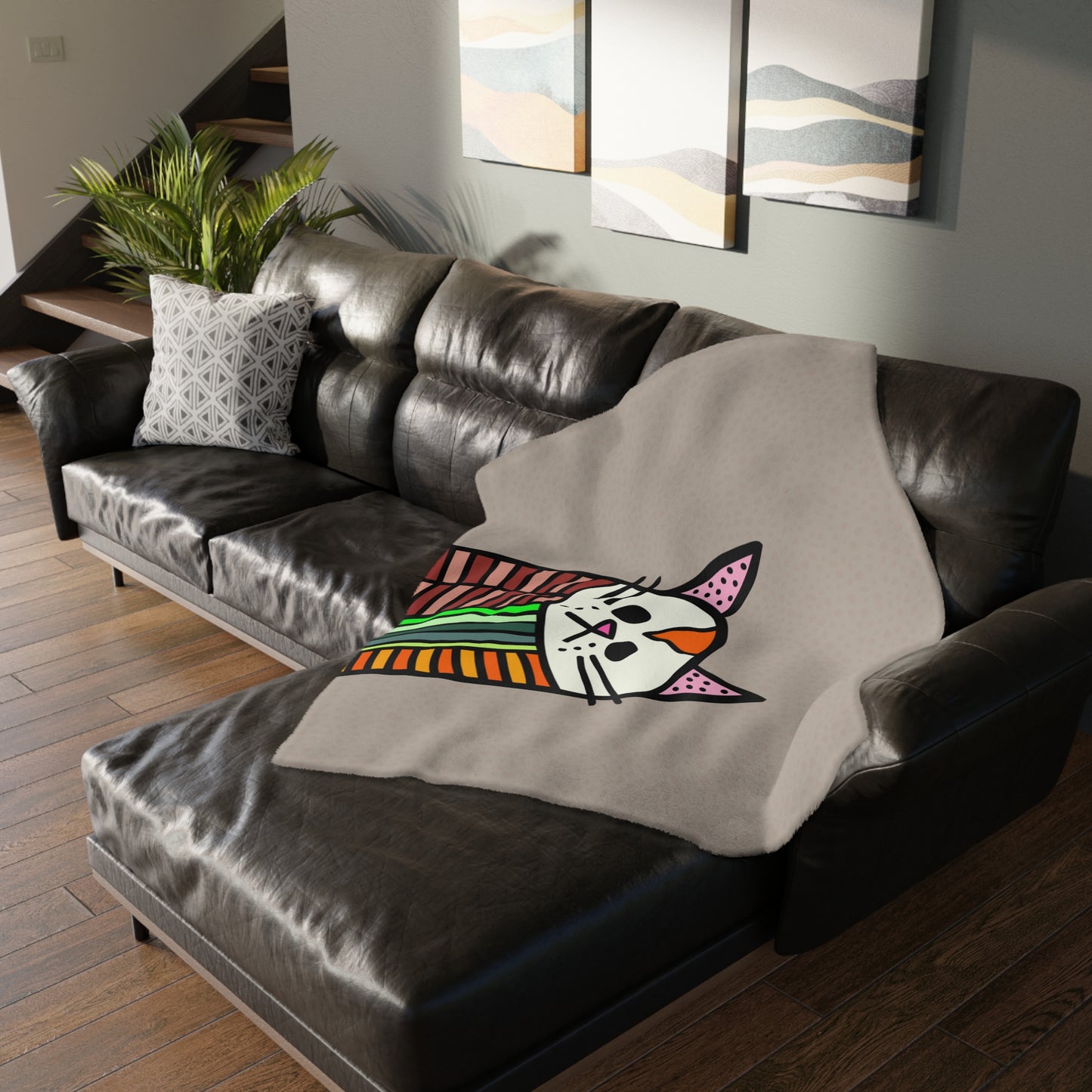 Gray blanket with a colorful cat design draped over a brown leather sofa in a living room.