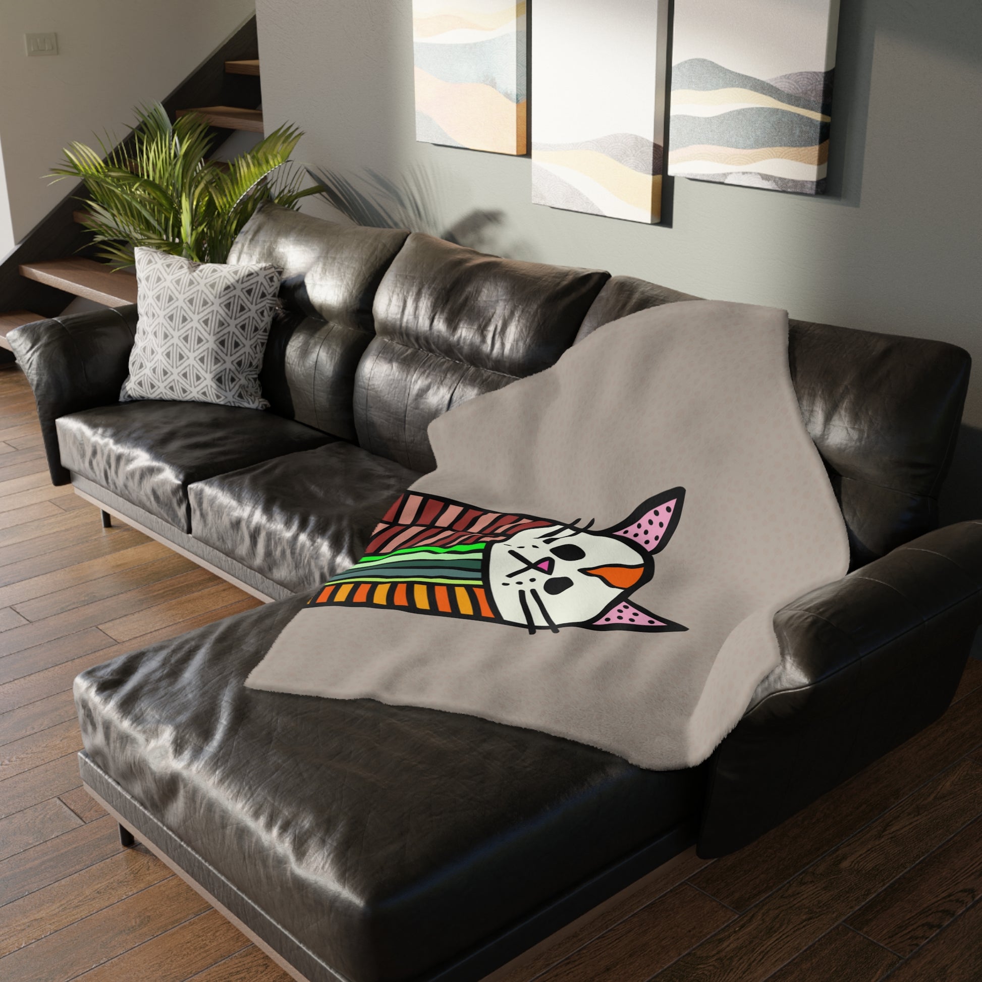 Gray blanket with a colorful cat design draped over a brown leather sofa in a living room.