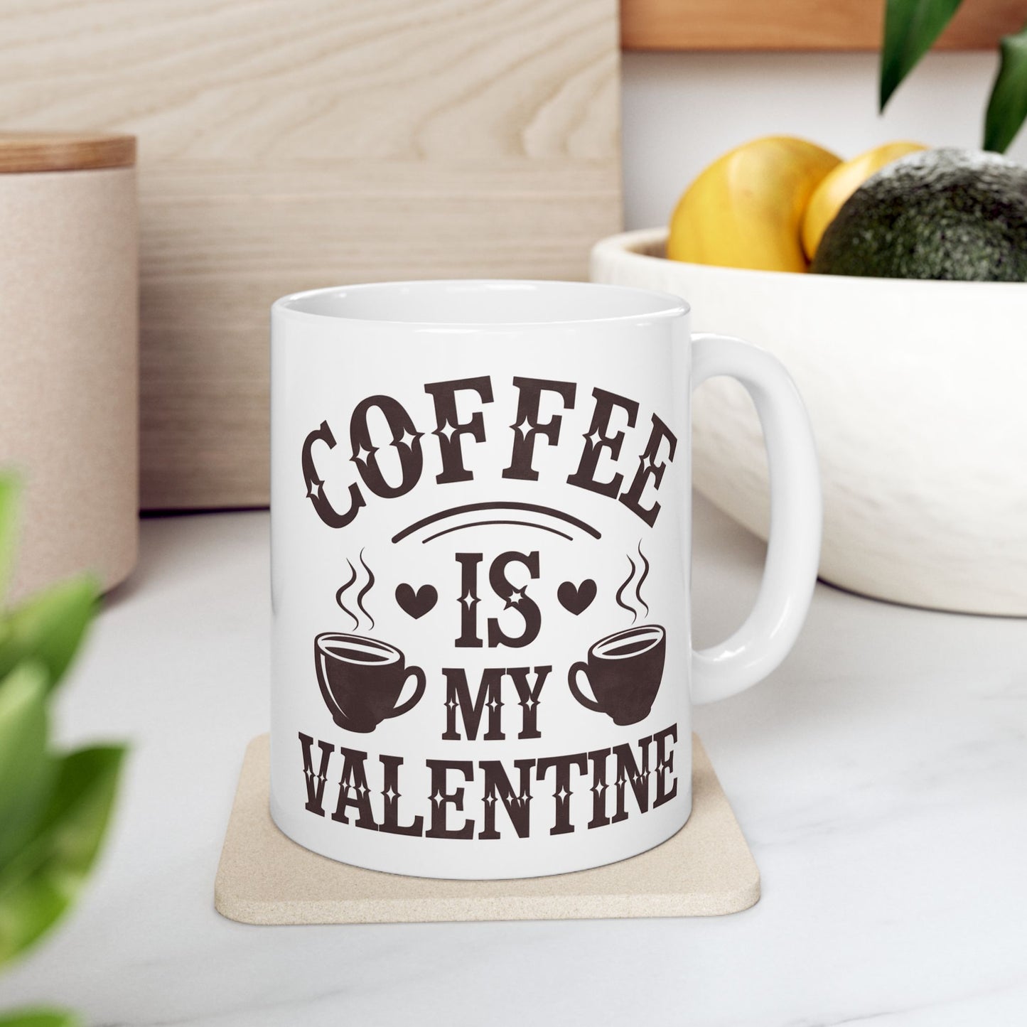 White mug with 'Coffee is my Valentine' text on a wooden coaster, surrounded by plants and a bowl of fruit.