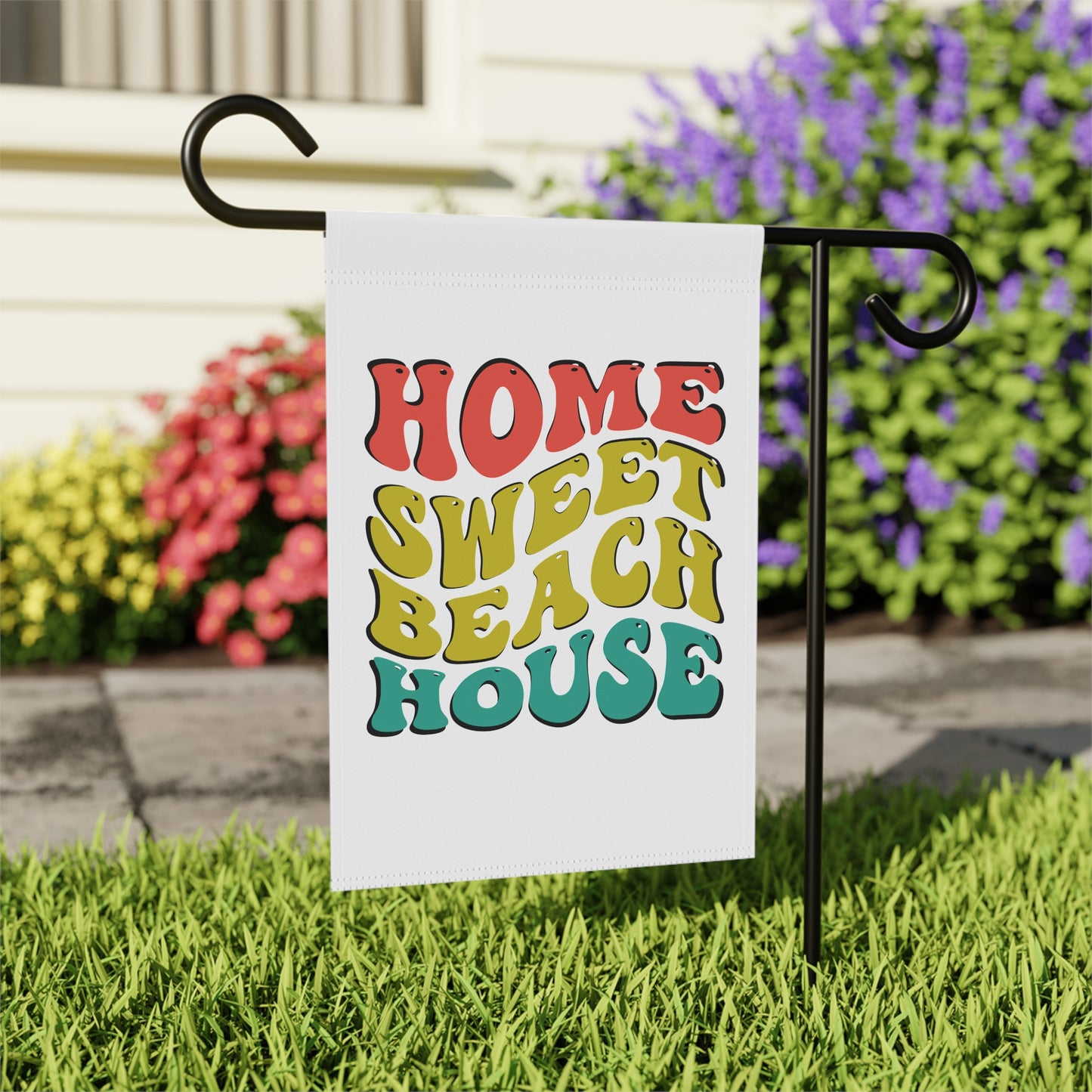 Garden flag with 'Home Sweet Beach House' text on a grassy lawn with flowers in the background