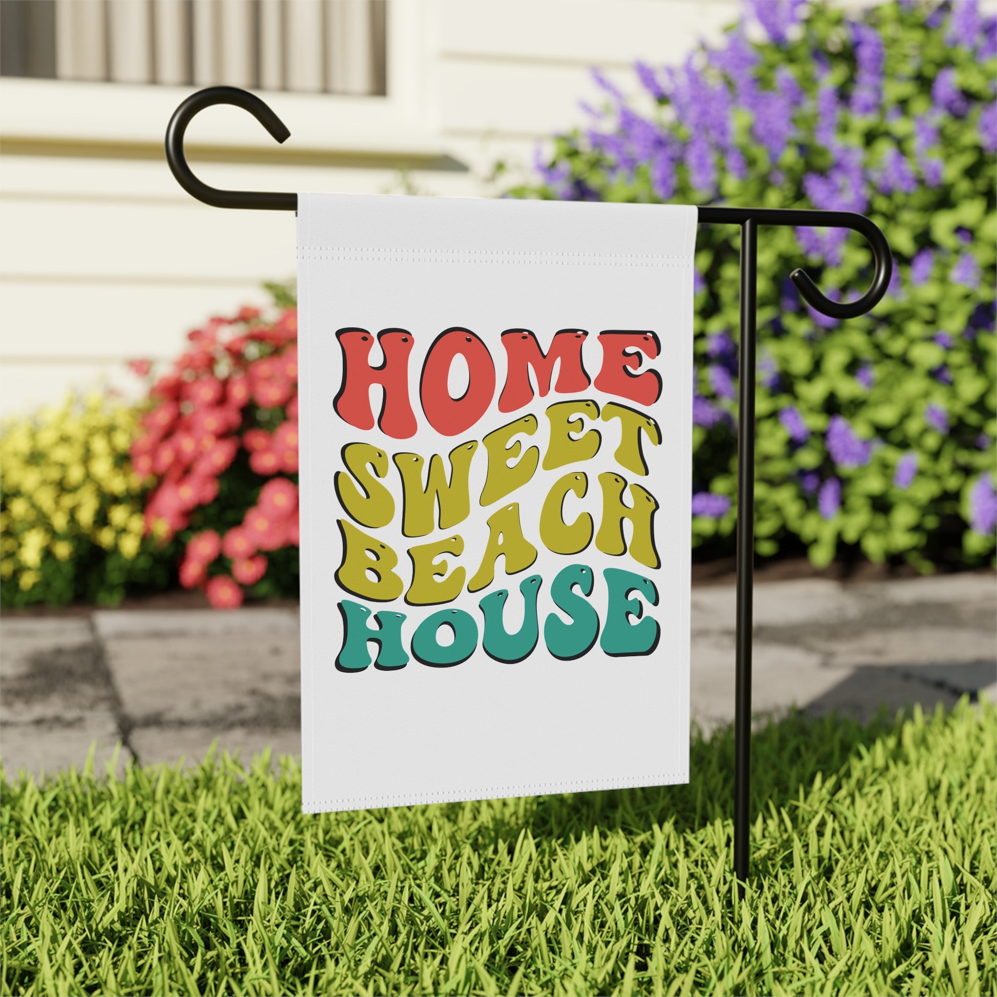 Garden flag with 'Home Sweet Beach House' text on a grassy lawn with flowers in the background