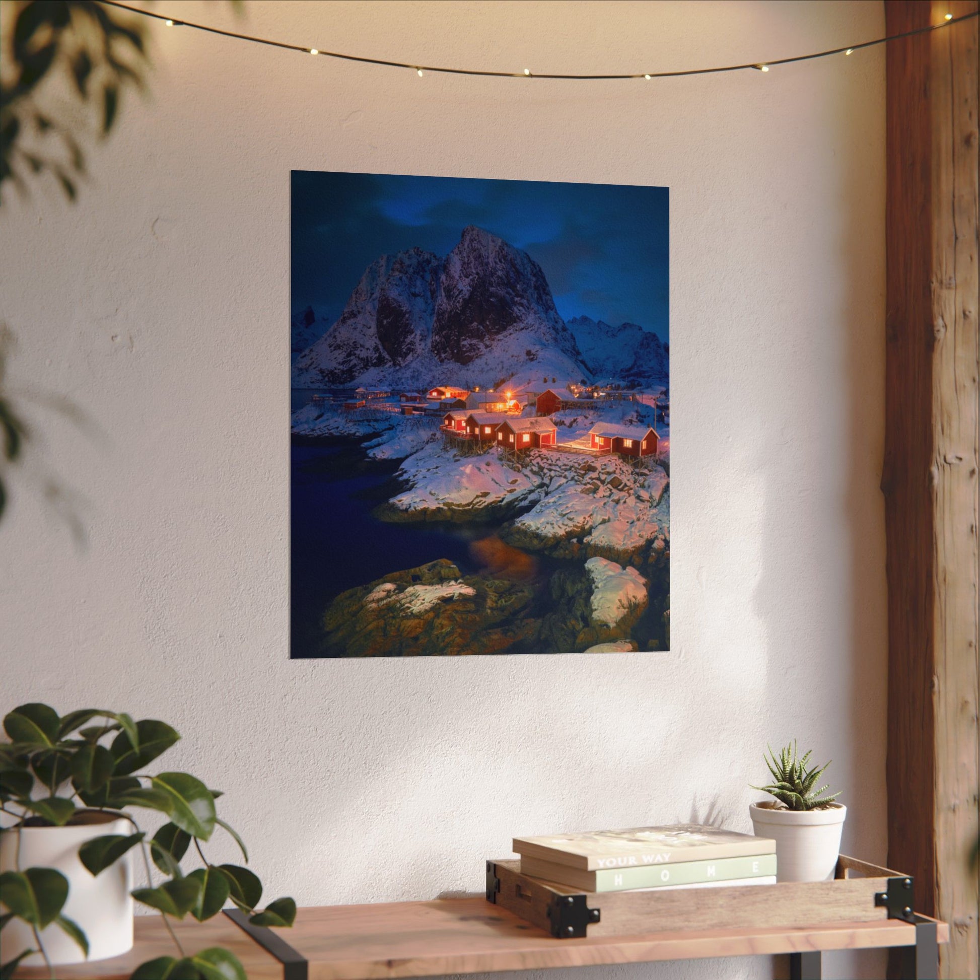 Framed artwork of a mountainous landscape with a village on a wall in a room with plants and a wooden table.