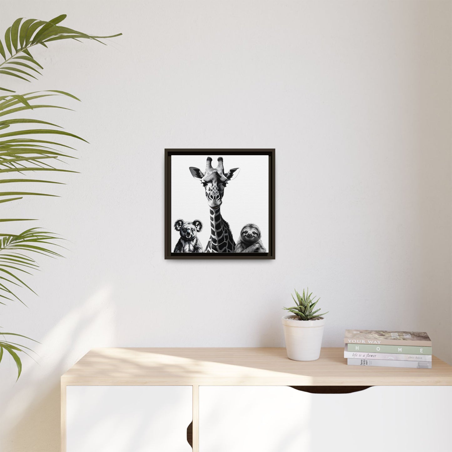 Framed artwork of a giraffe and sloths on a wall above a desk with a plant and books.