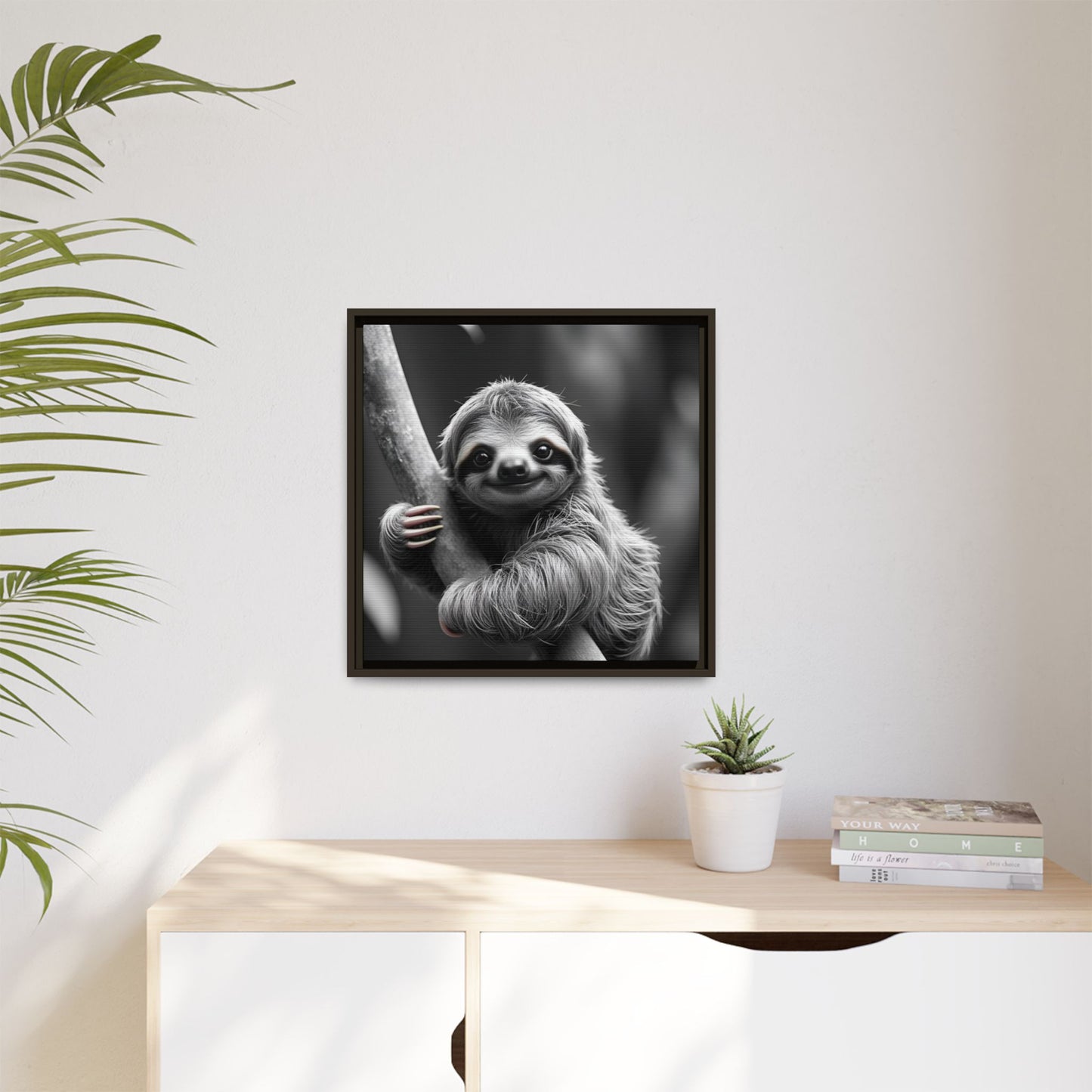 Framed black and white photo of a sloth on a wall above a desk with a plant and books.