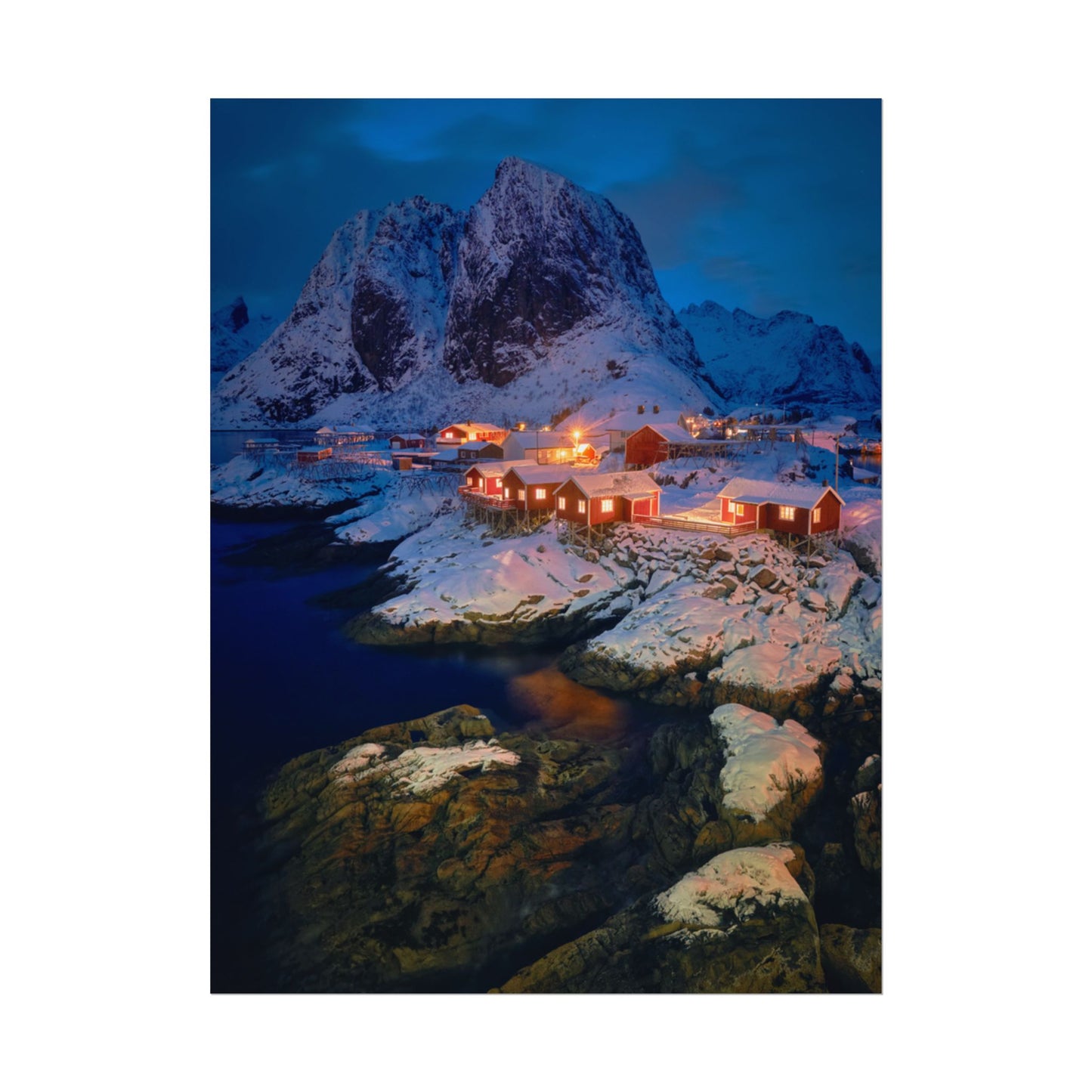 Snowy mountain landscape with illuminated buildings and a body of water.