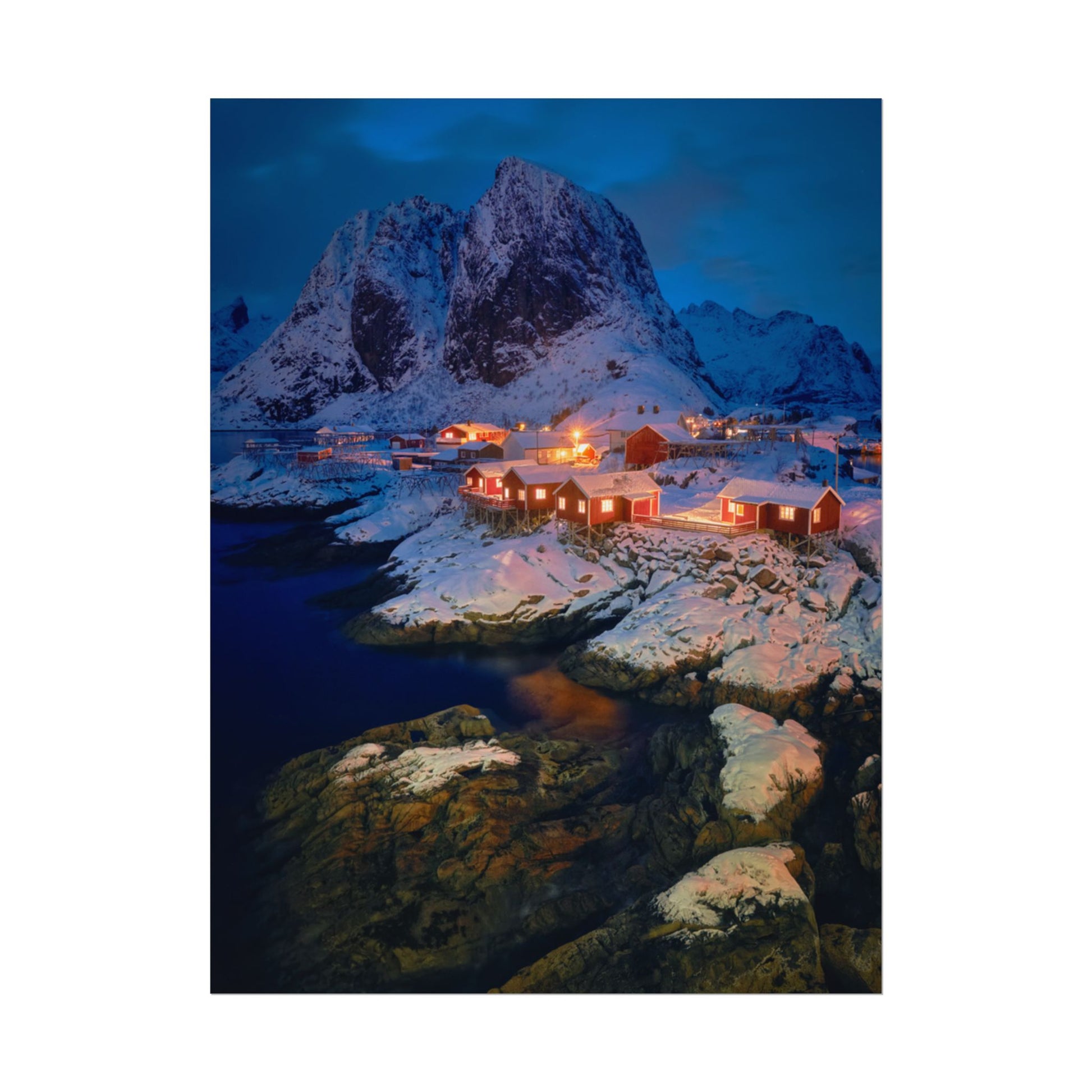 Snowy mountain landscape with illuminated buildings and a body of water.