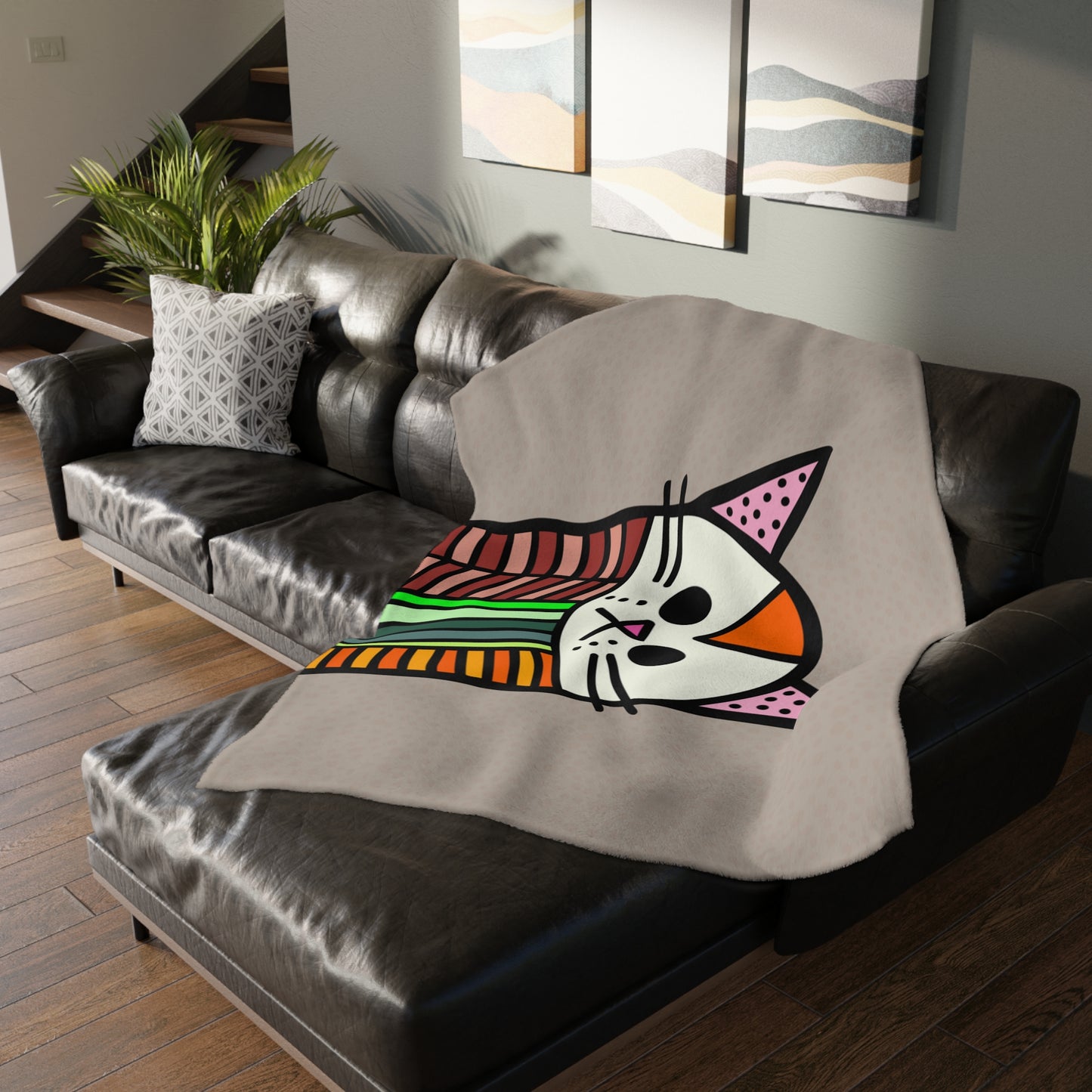 Gray blanket with a colorful cat design draped over a black leather couch in a living room.