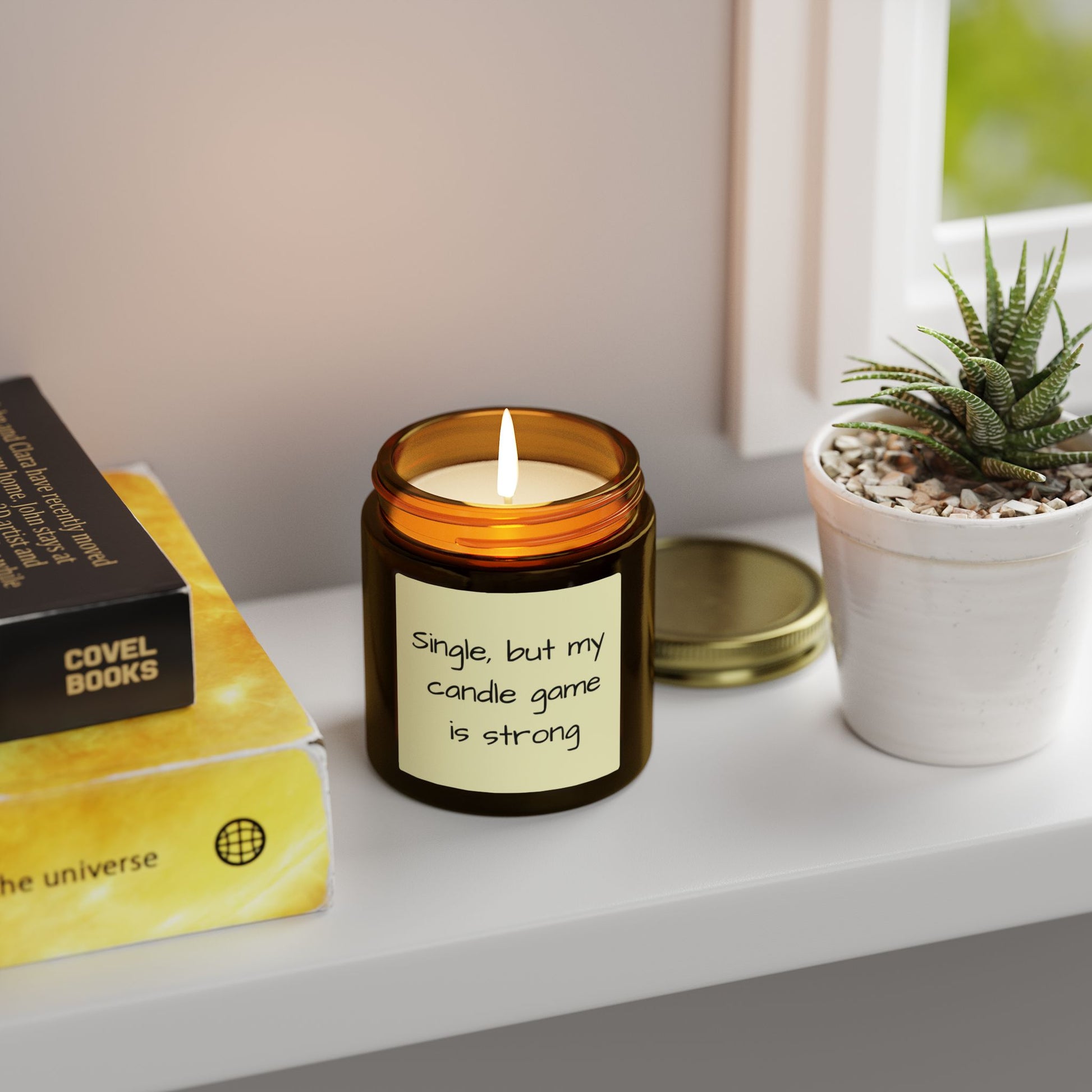 Candle with a humorous label on a shelf next to a book and plant