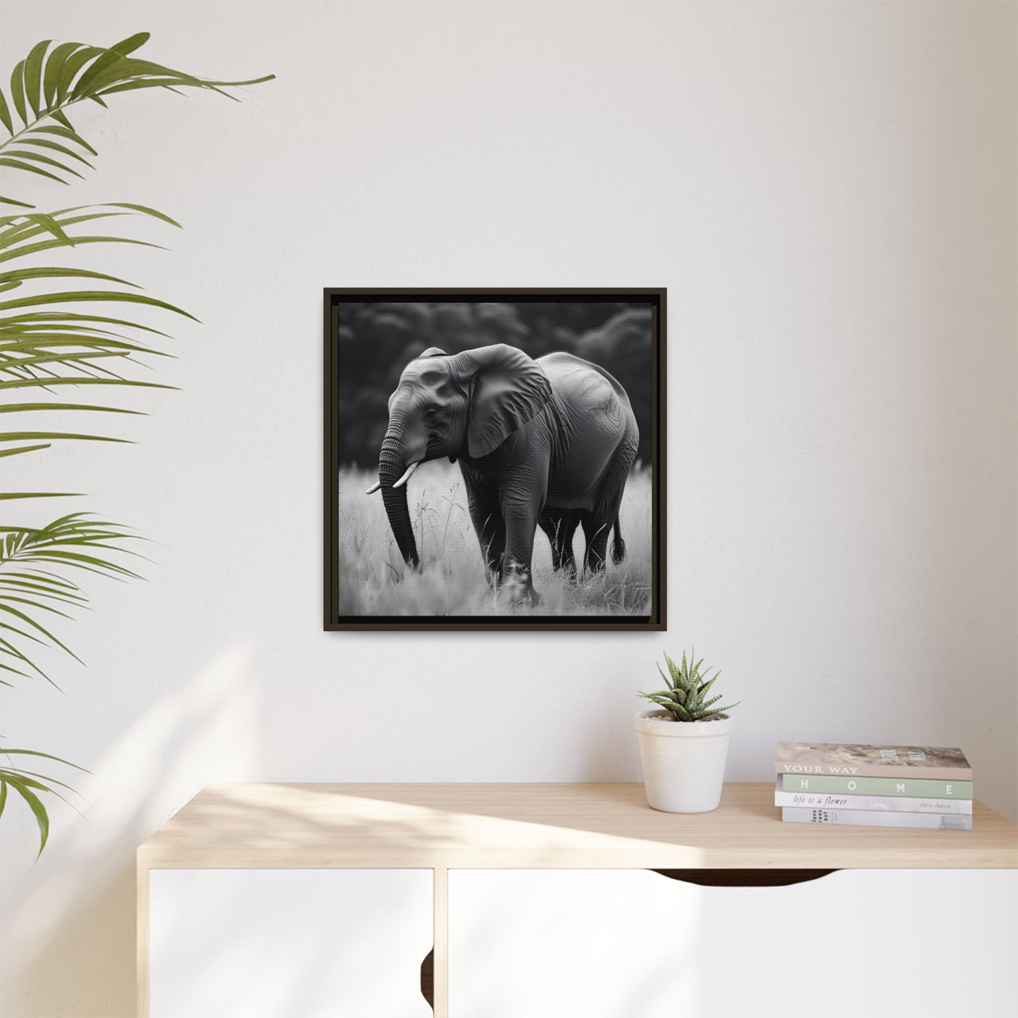 Framed black and white elephant photograph on a wall above a desk with a plant and books.
