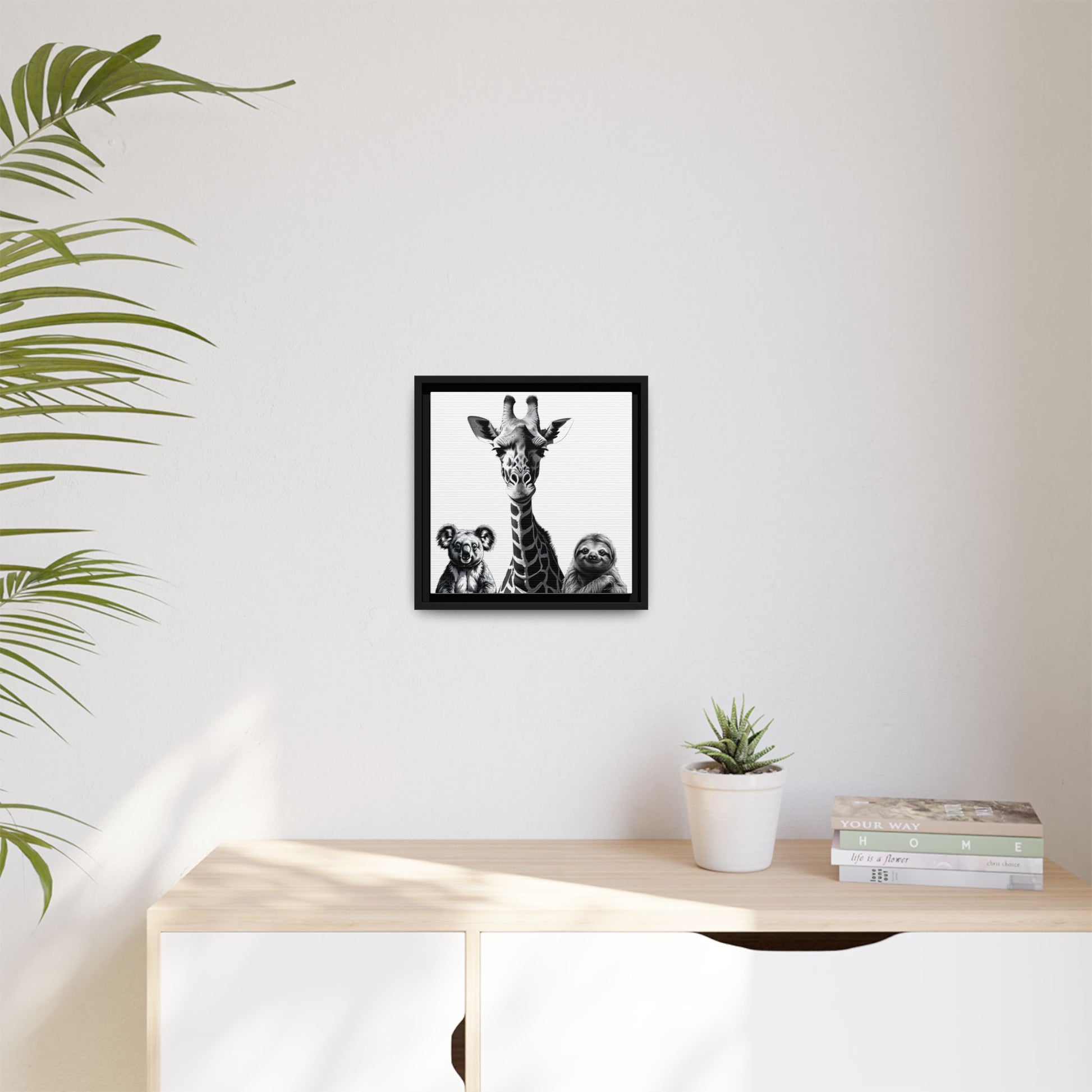 Framed artwork of a giraffe and sloths on a wall above a desk with a plant and books.