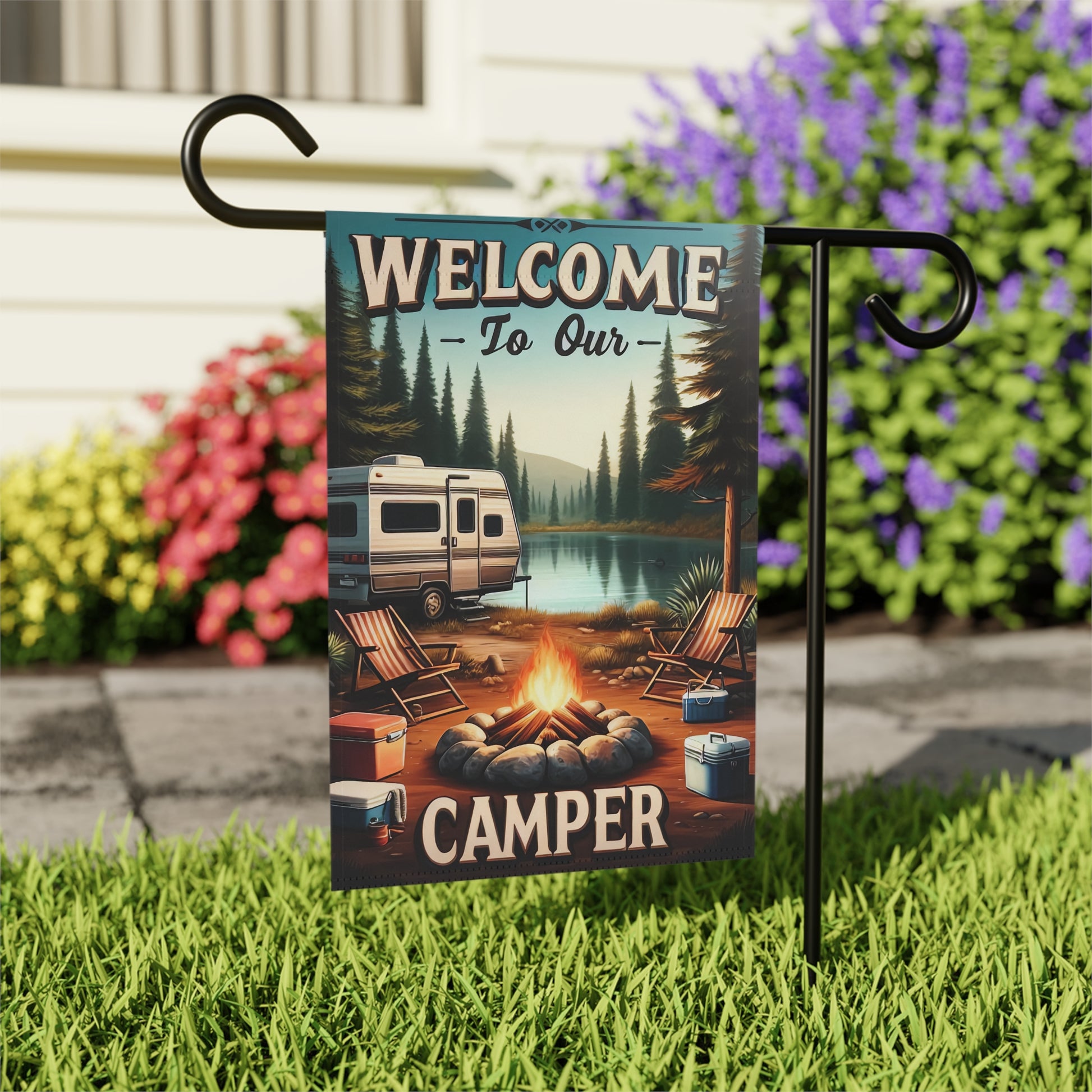 Garden flag with camping scene and 'Welcome to Our Camper' text, attached to a stand with flowers in the background.