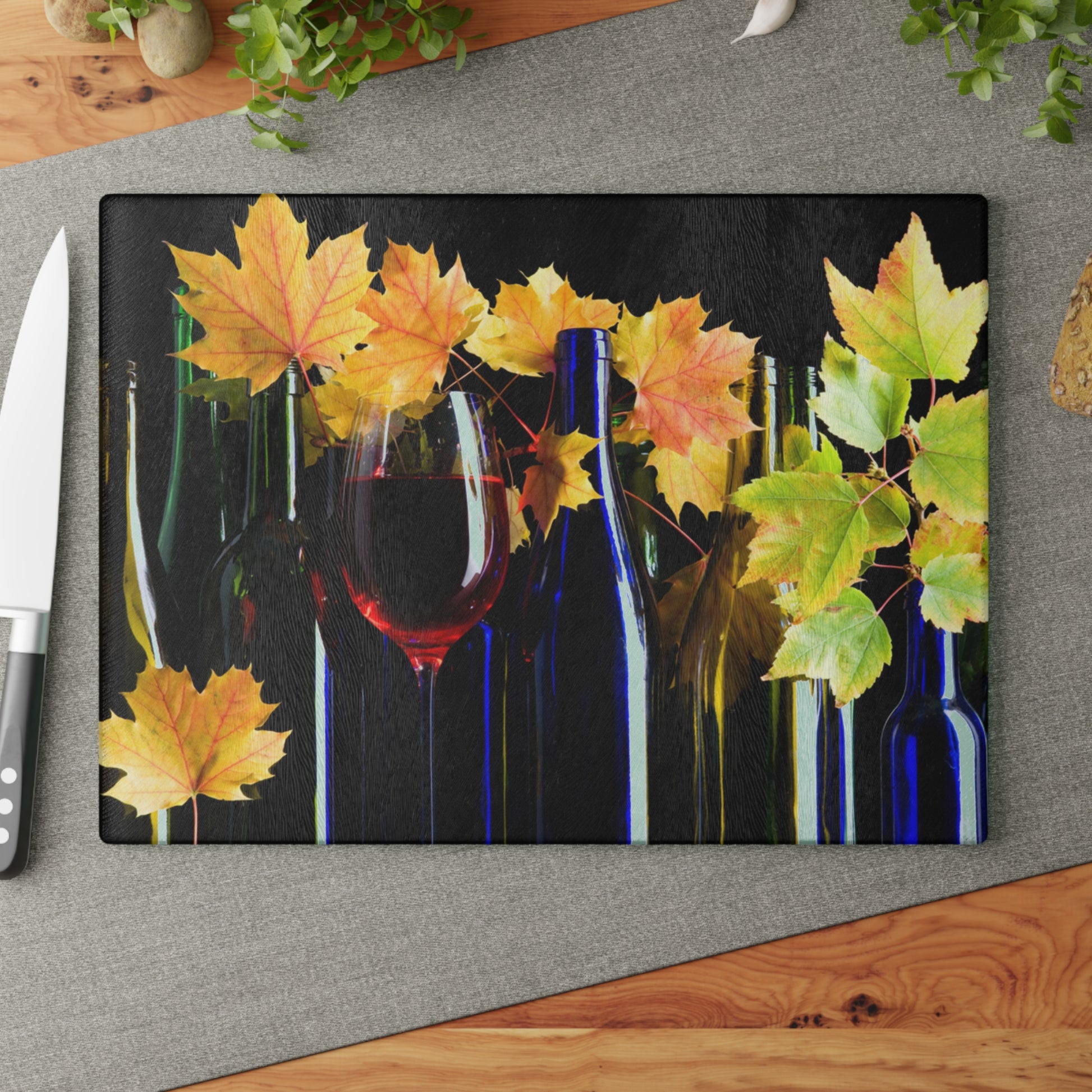 Decorative cutting board with autumn leaves, wine glasses, and bottles on a gray surface.