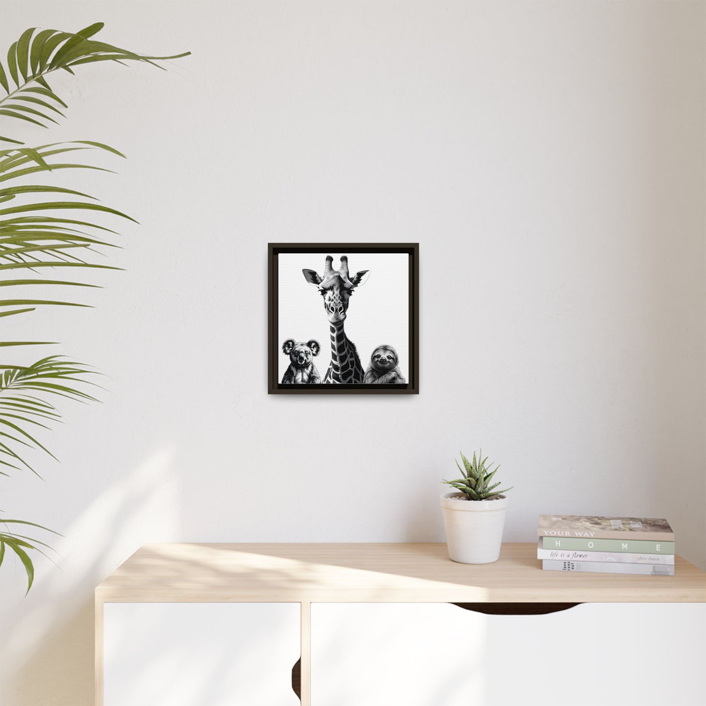 Framed artwork of a giraffe and sloths on a wall above a desk with a plant and books.