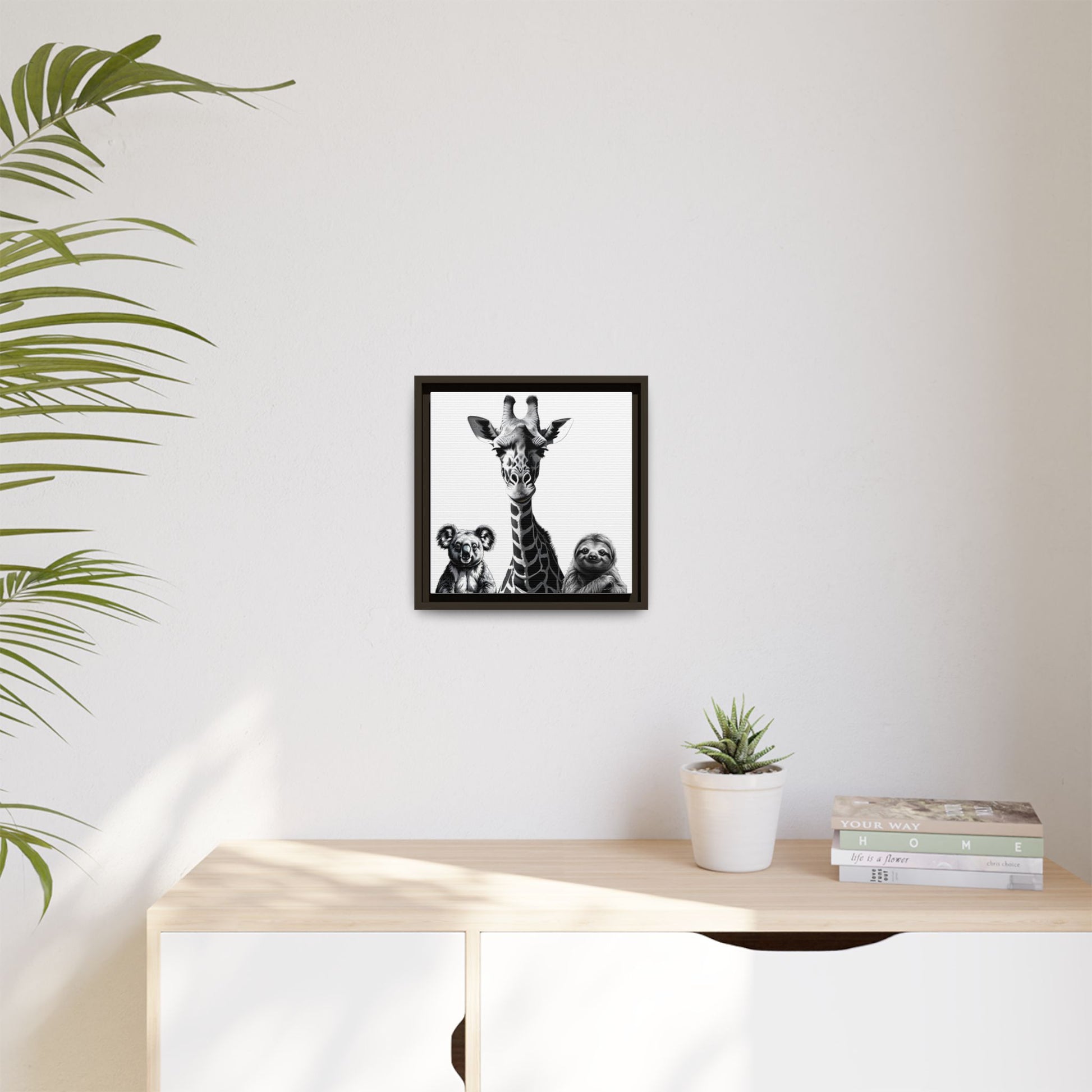 Framed artwork of a giraffe and sloths on a wall above a desk with a plant and books.