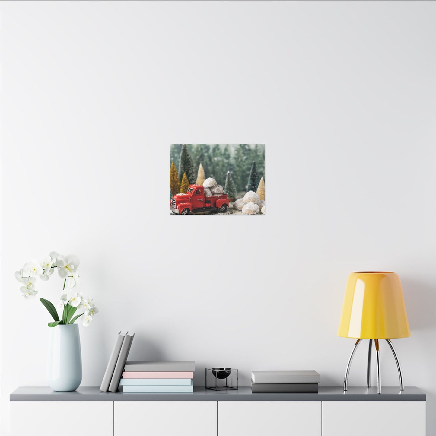 Decorative wall art of a red truck and pine trees on a white wall above a shelf with a lamp and books.