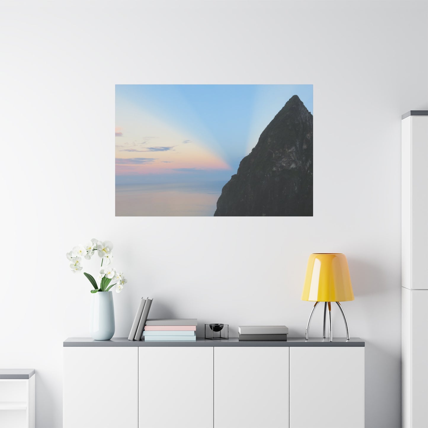 Framed artwork of a mountain landscape on a wall above a white cabinet with a yellow lamp.