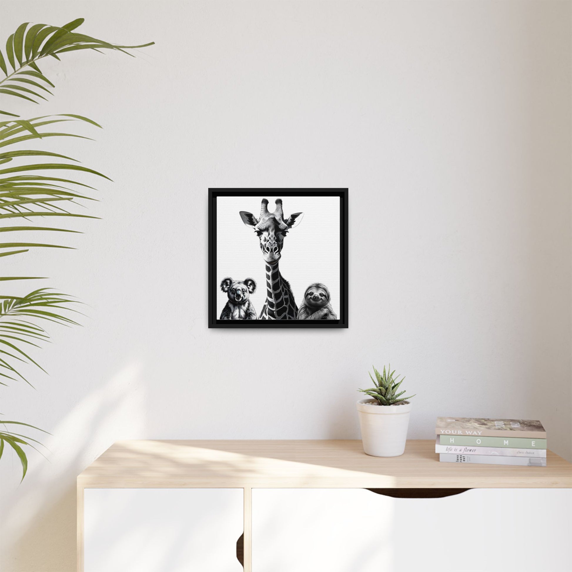Framed artwork of a giraffe and sloths on a wall above a desk with a plant and books.