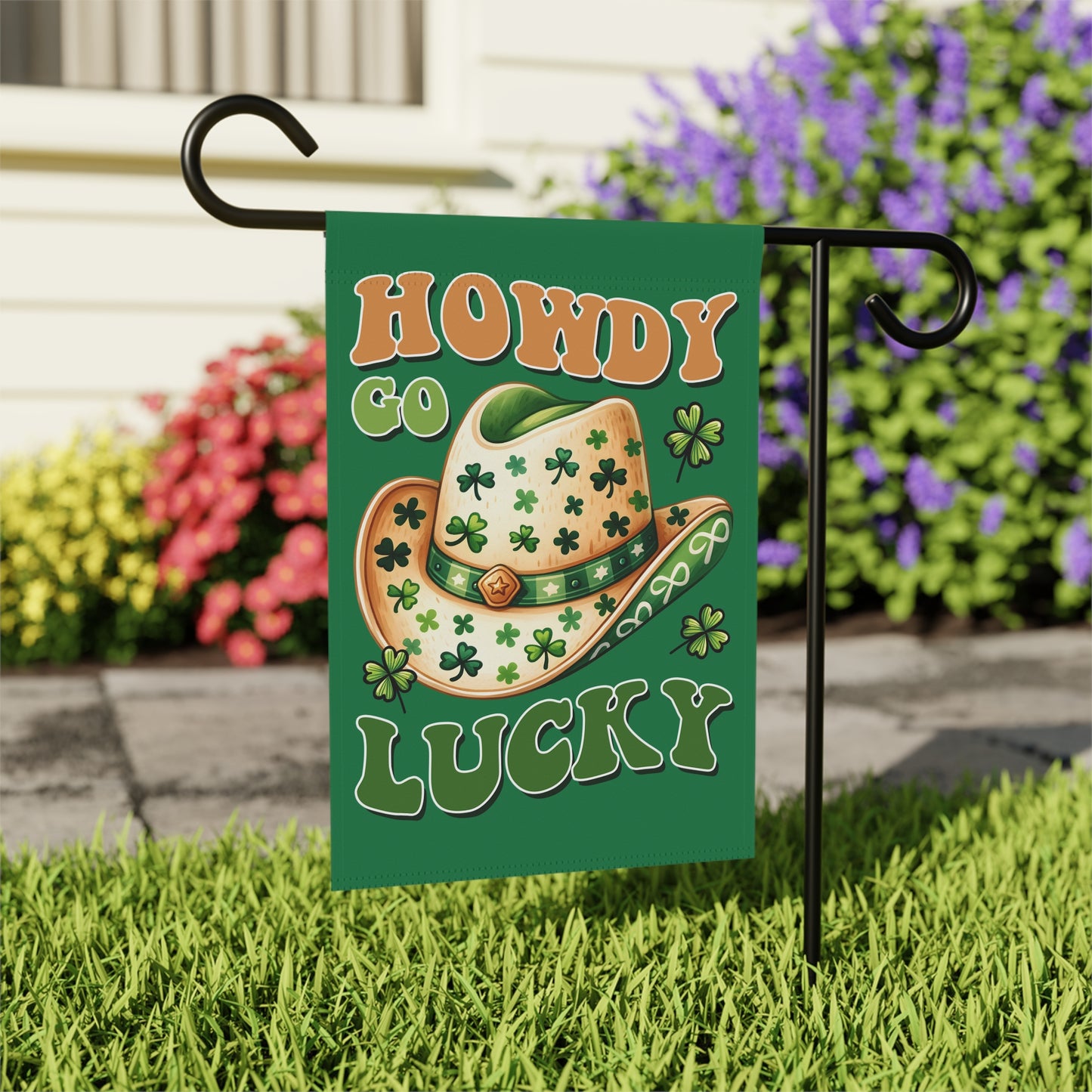 Decorative garden flag with a cowboy hat and shamrocks on a grassy area with flowers in the background.