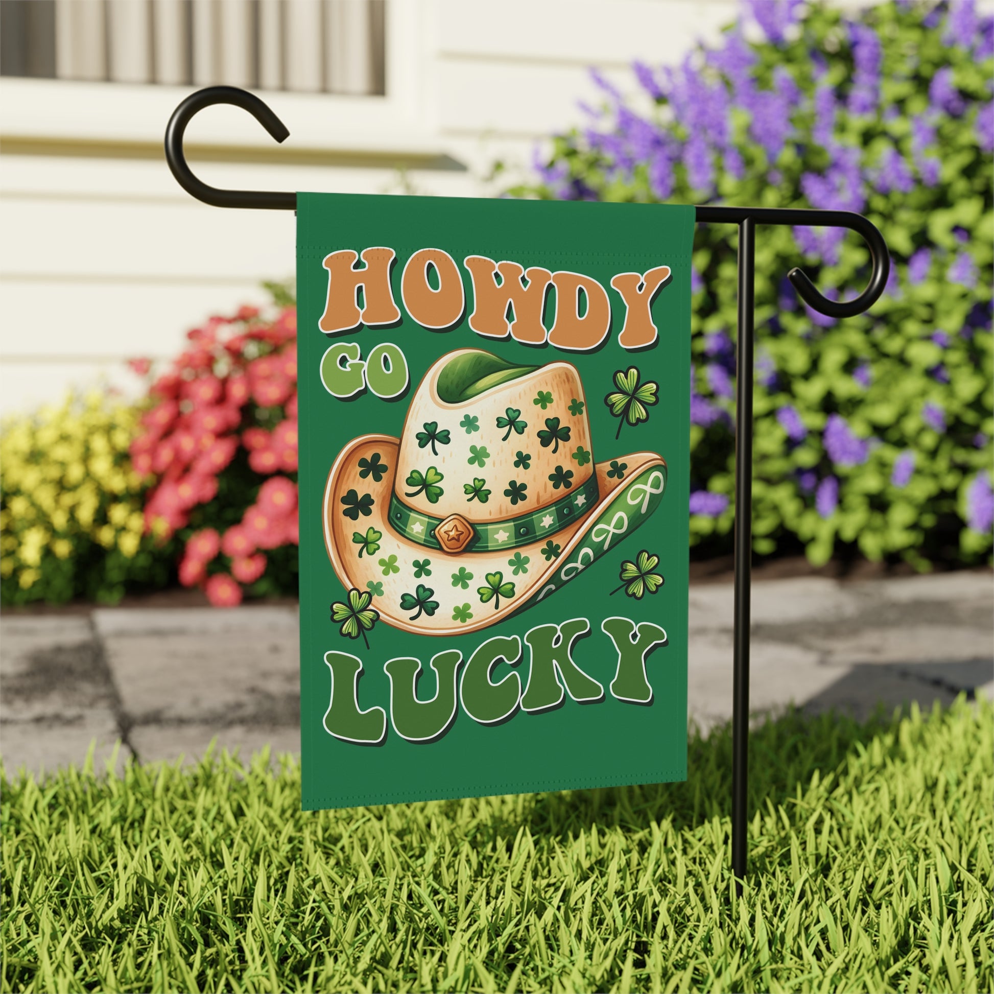 Decorative garden flag with a cowboy hat and shamrocks on a grassy area with flowers in the background.