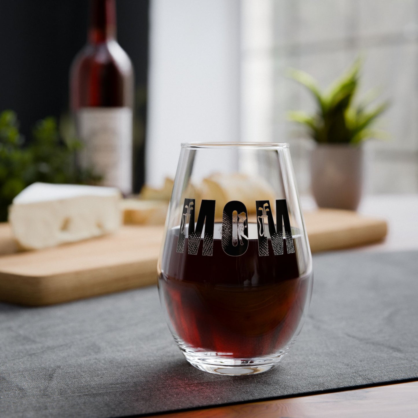 Glass of red wine with 'MOM' engraving on a table with cheese and wine bottle in the background.