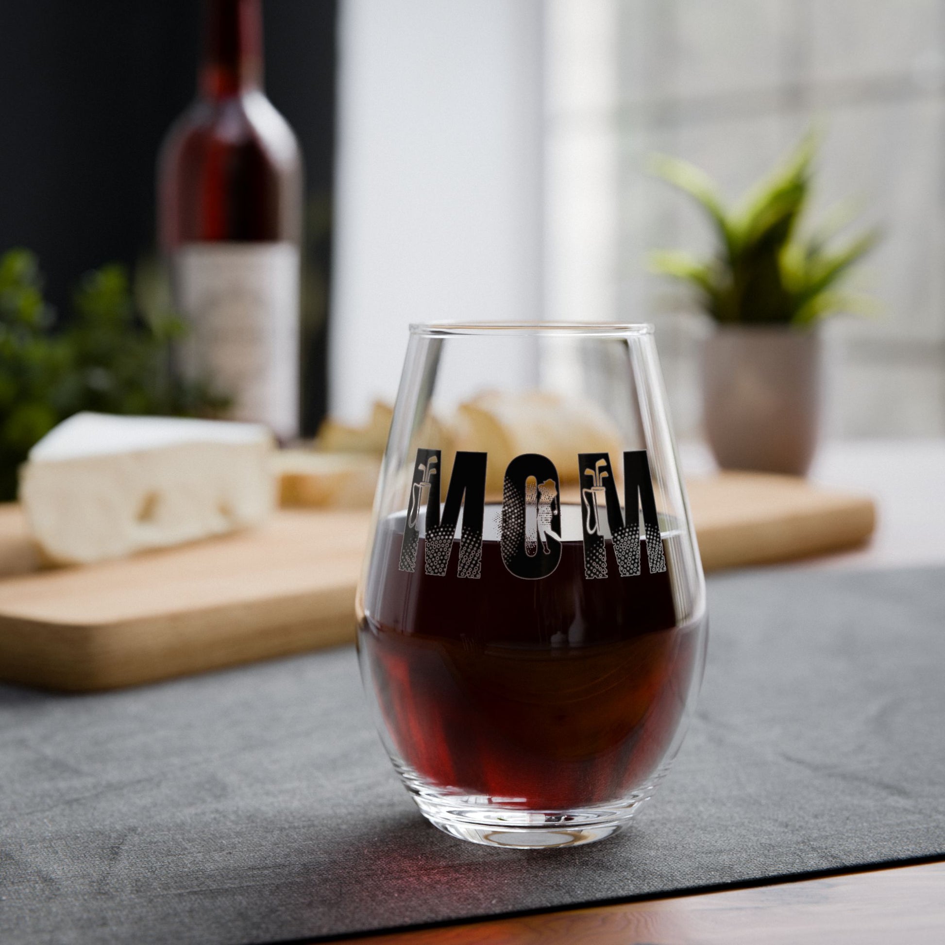 Glass of red wine with 'MOM' engraving on a table with cheese and wine bottle in the background.