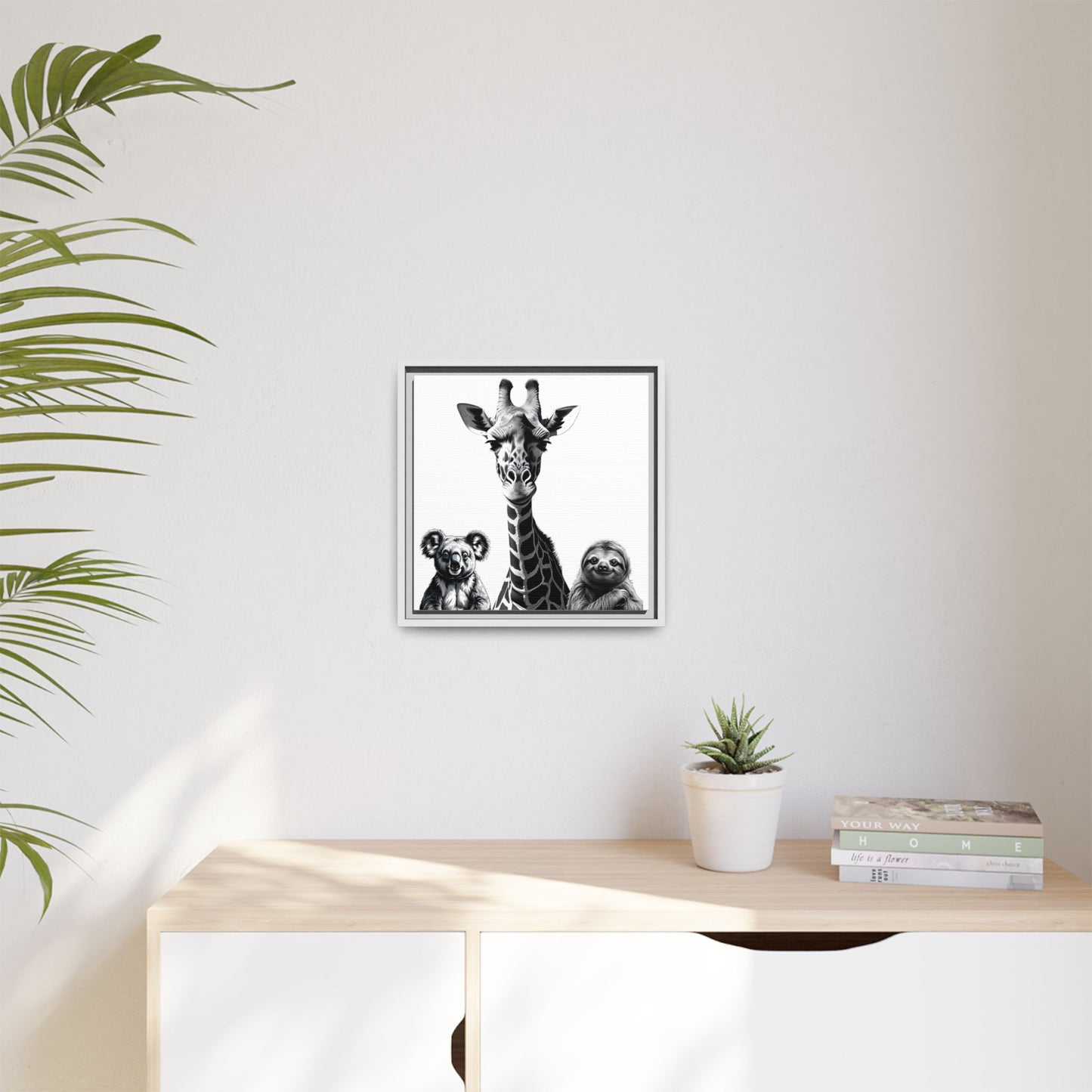 Framed artwork of a giraffe and sloths on a wall above a desk with a plant and books.