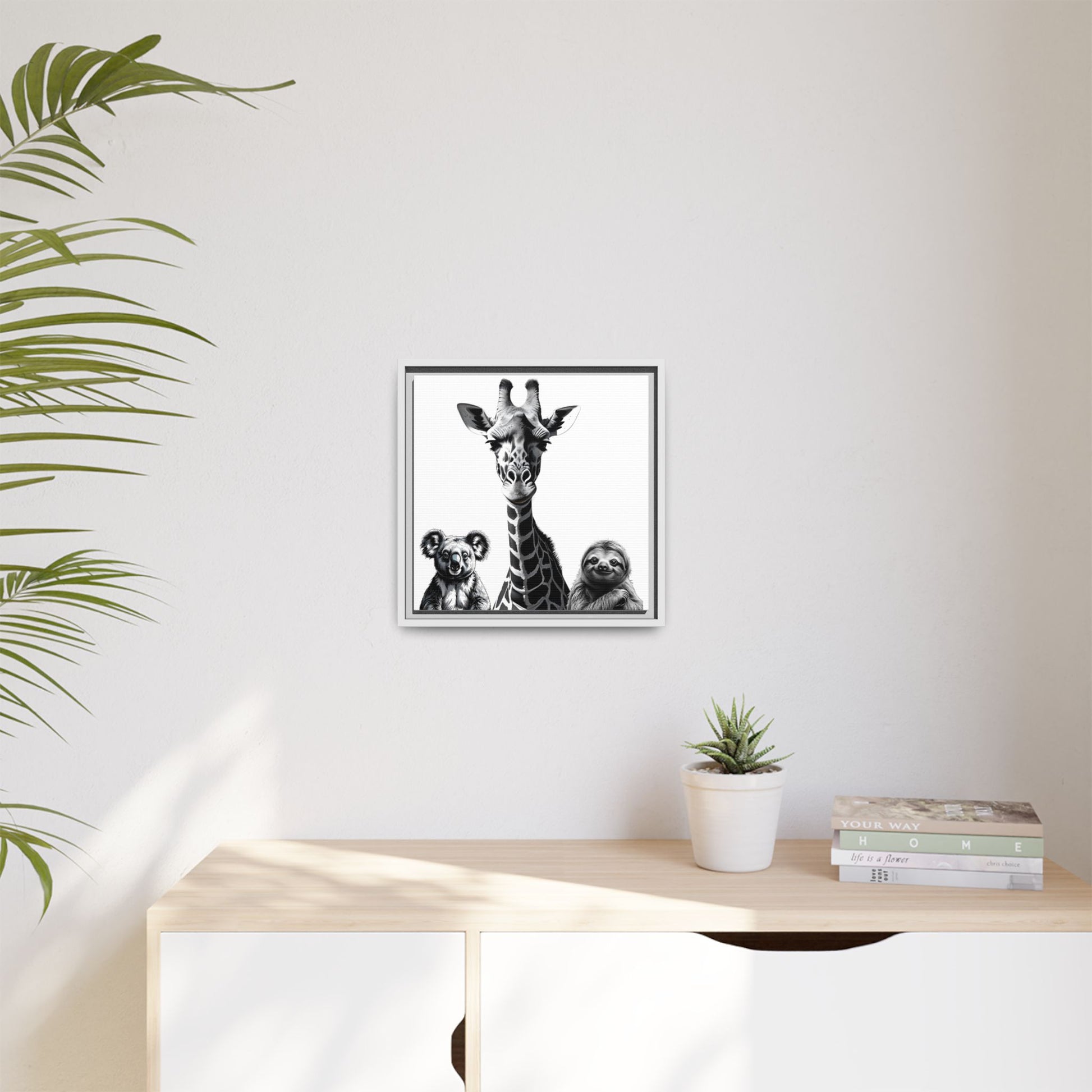 Framed artwork of a giraffe and sloths on a wall above a desk with a plant and books.