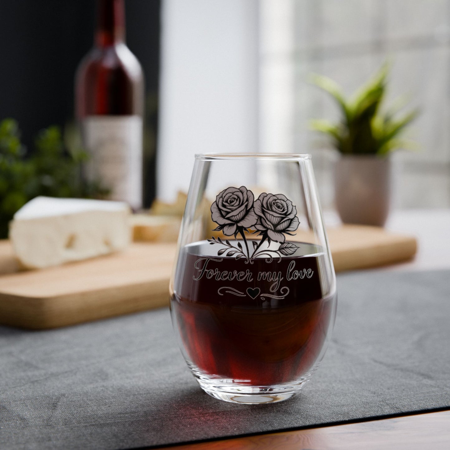 Stemless wine glass with engraved roses and 'Forever my love' text, filled with red wine, on a table.