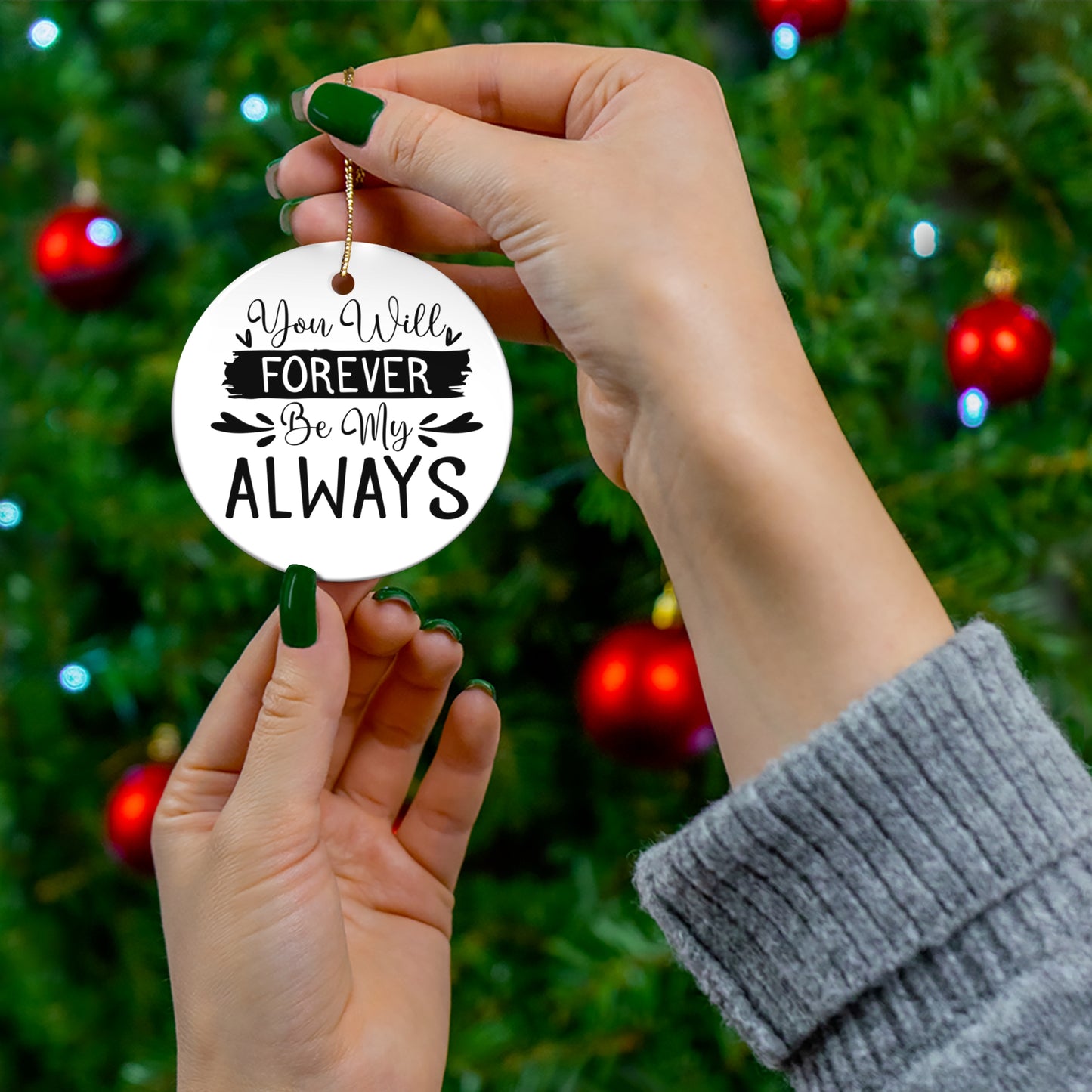 Hand holding a decorative ornament with 'You will forever be my always' text against a Christmas tree background.