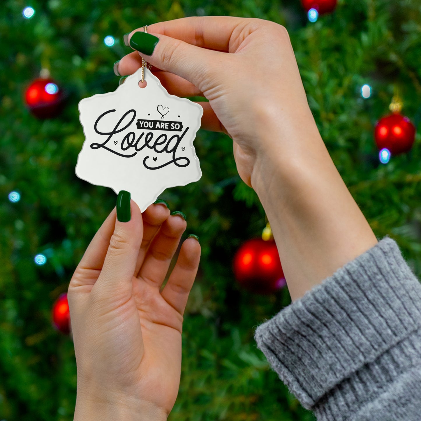 Hand holding a decorative ornament with 'You are so loved' text against a Christmas tree background.