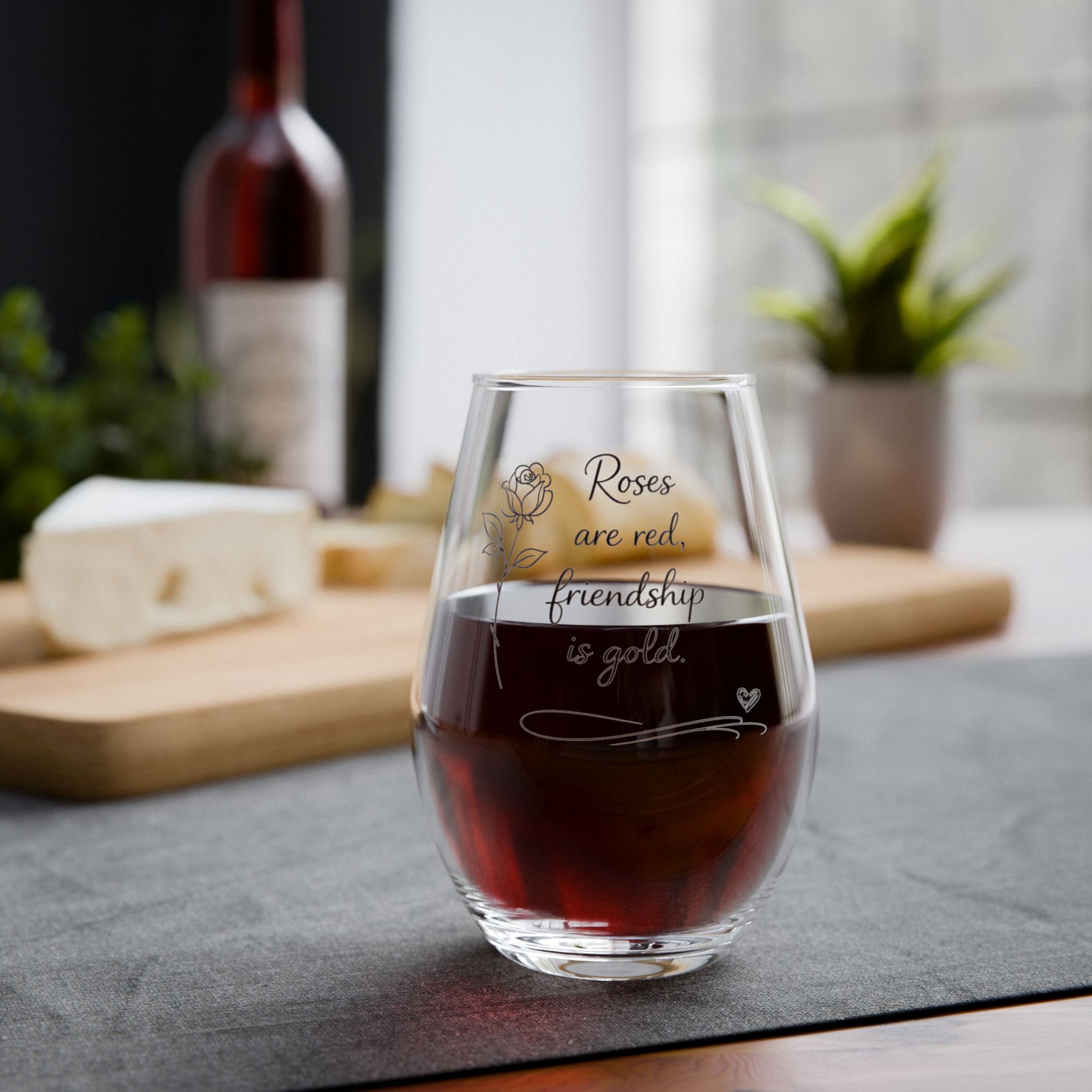 Stemless wine glass with engraved message on a table with a bottle of wine and cheese in the background.