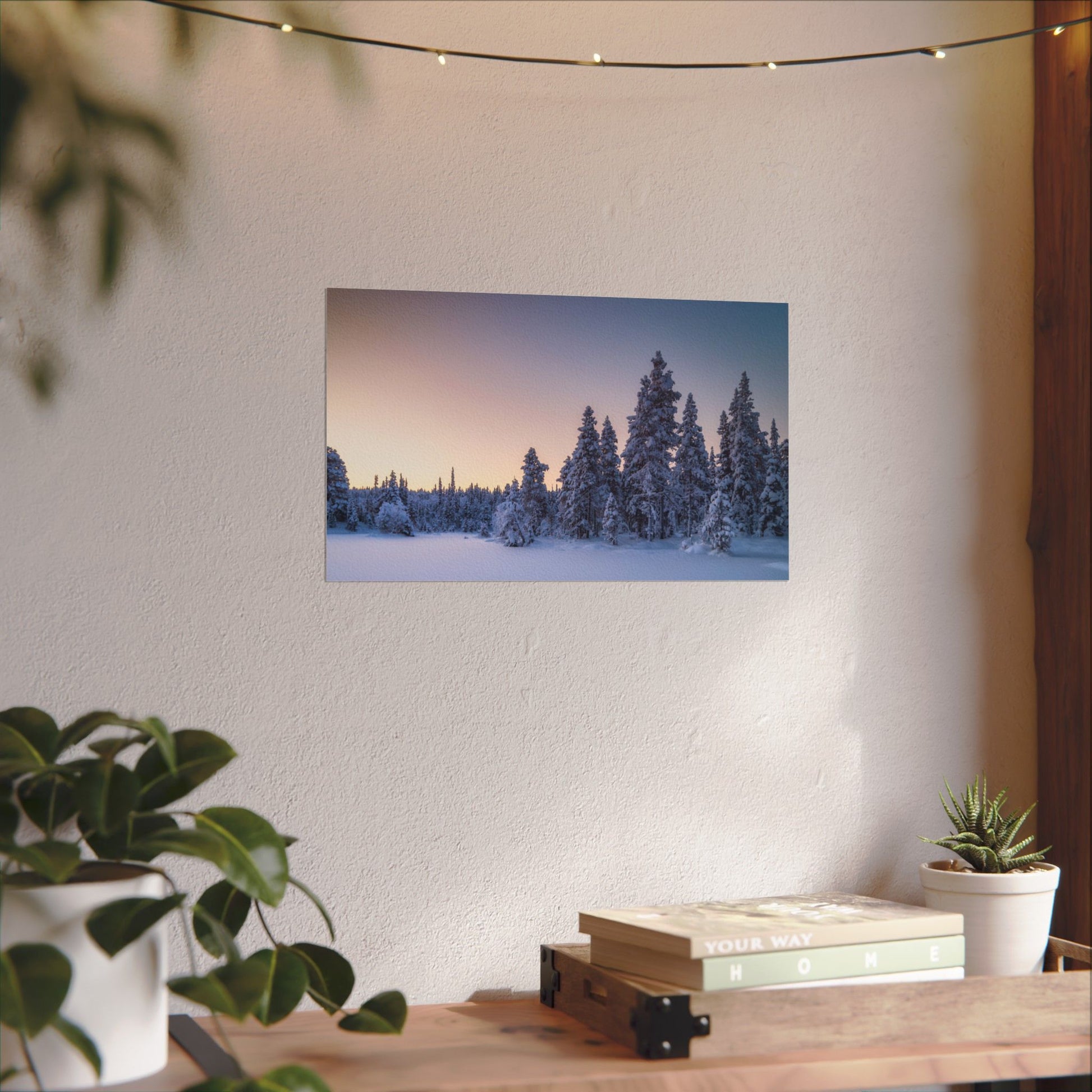 Framed winter landscape print on a wall above a wooden table with plants.