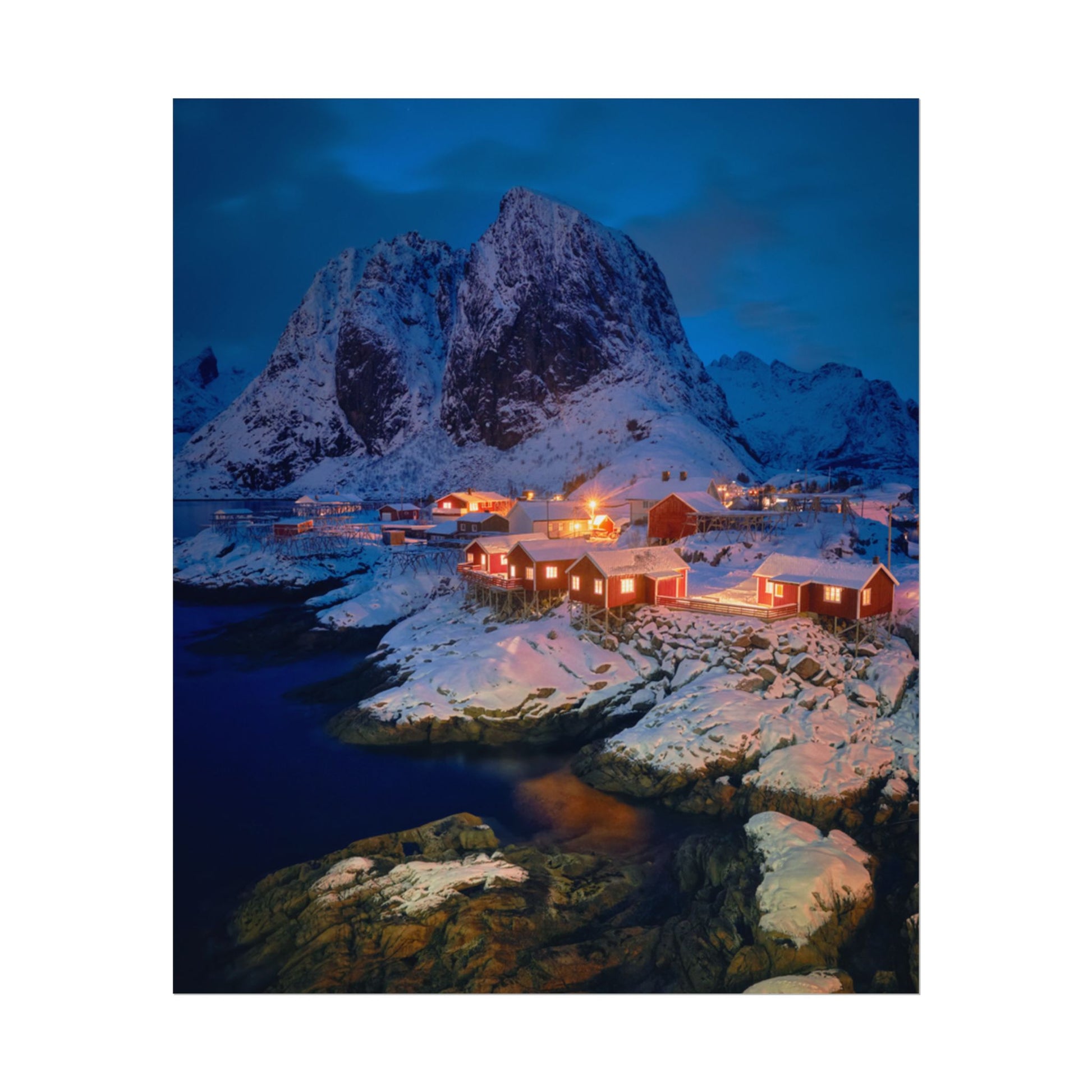 Snowy mountain landscape with illuminated cabins near a body of water at night.