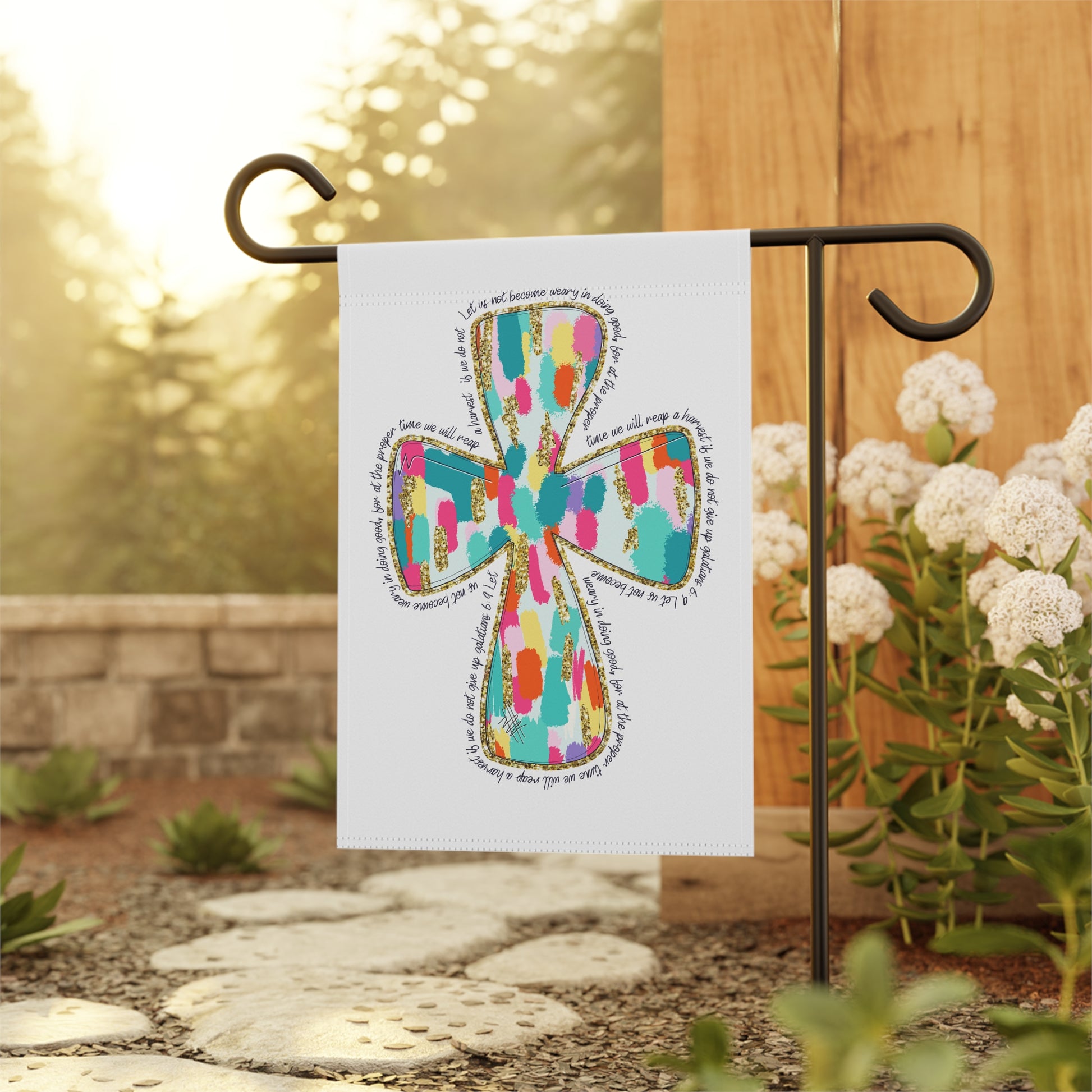 Colorful cross design on a garden flag with a blurred background of flowers and stones.