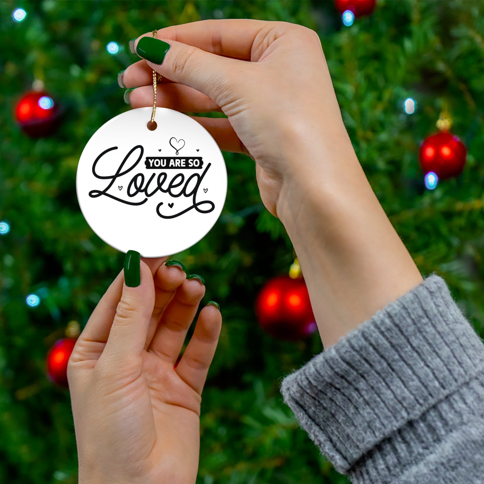 Hand holding a 'You are so loved' ornament with a Christmas tree background
