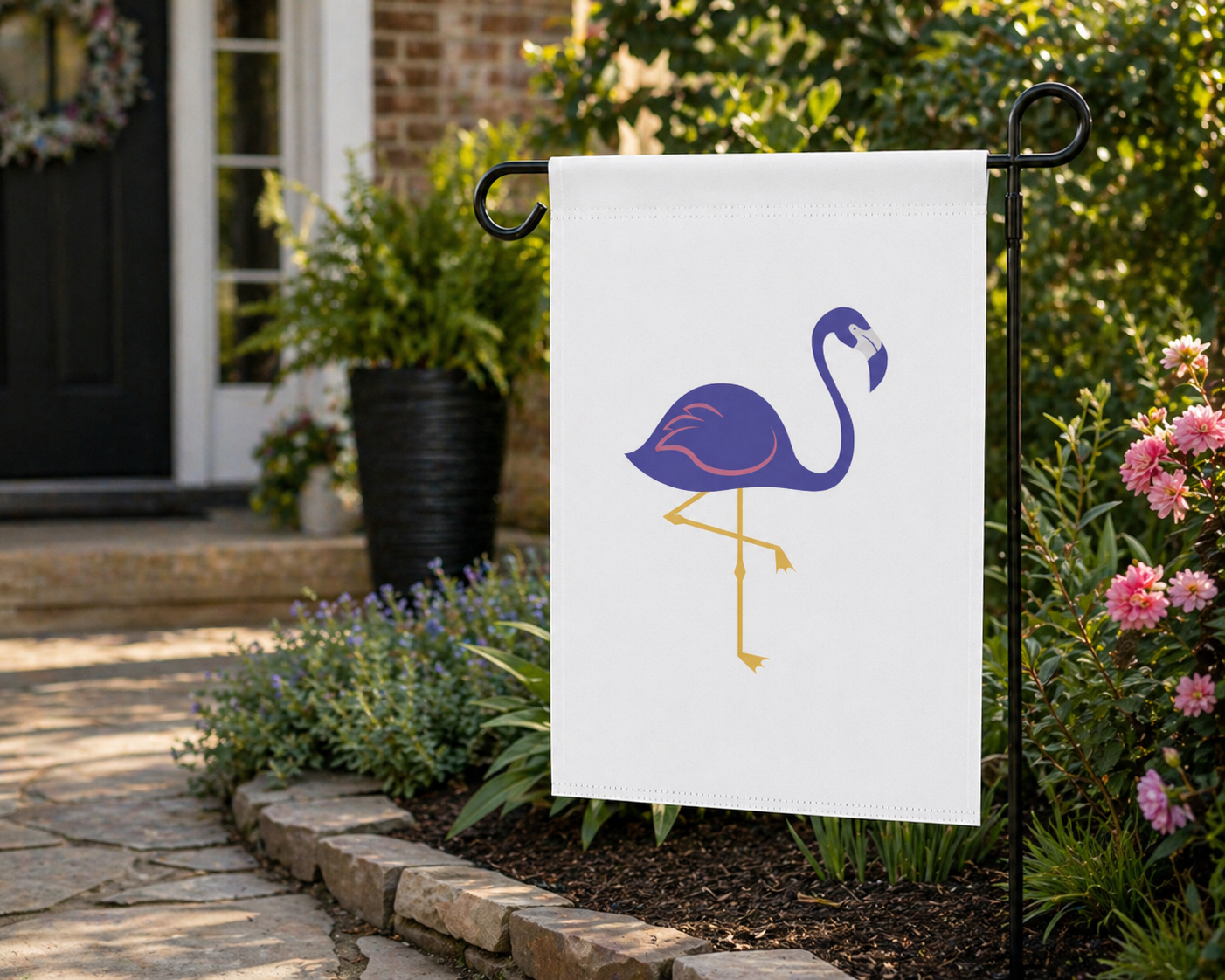 Flamingo-themed garden flag on a pole in front of a house with flowers and plants.